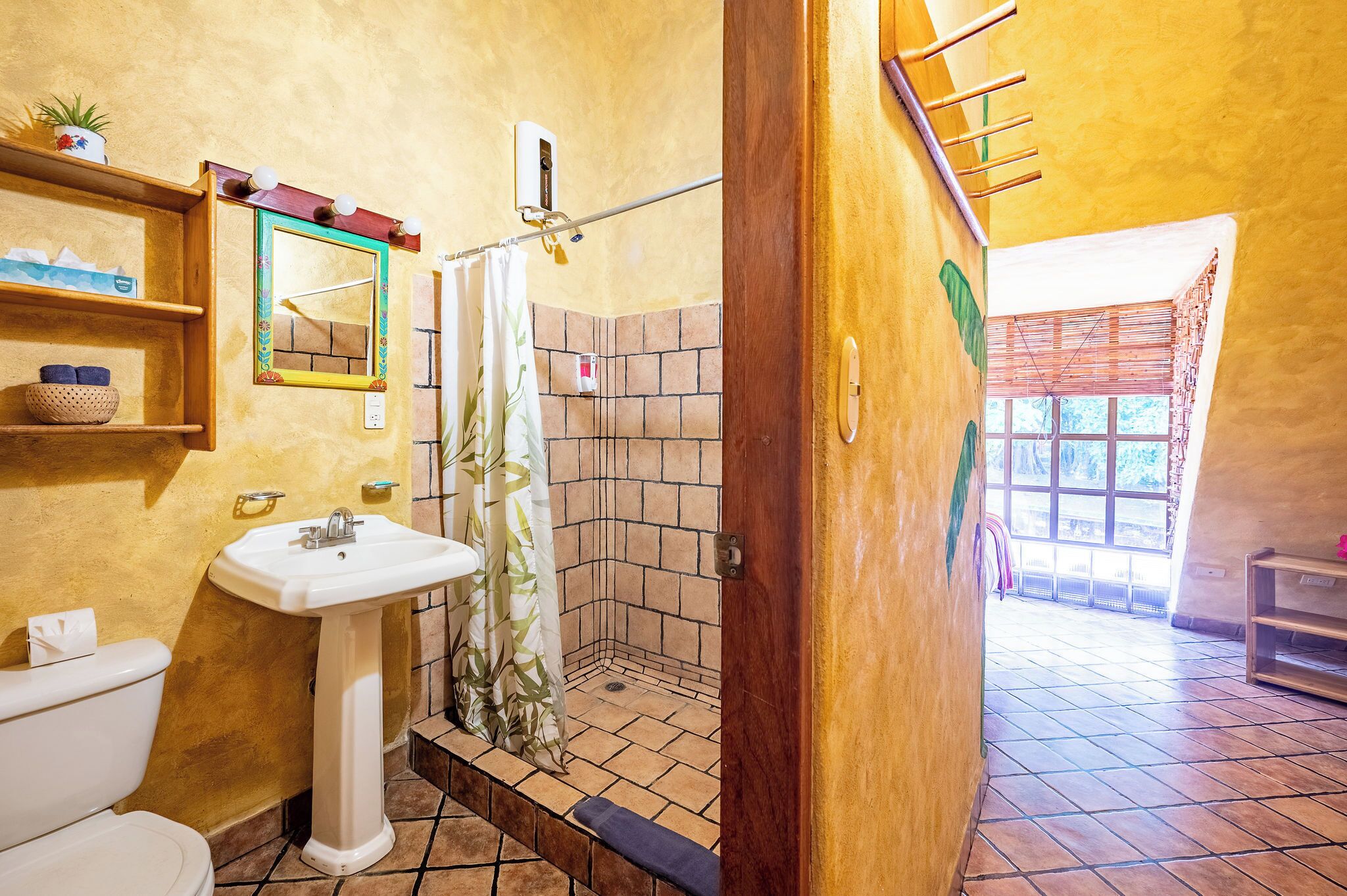 A bathroom with a white sink, a toilet, a shower with a tiled wall, and a wooden door leading to a room with a window.