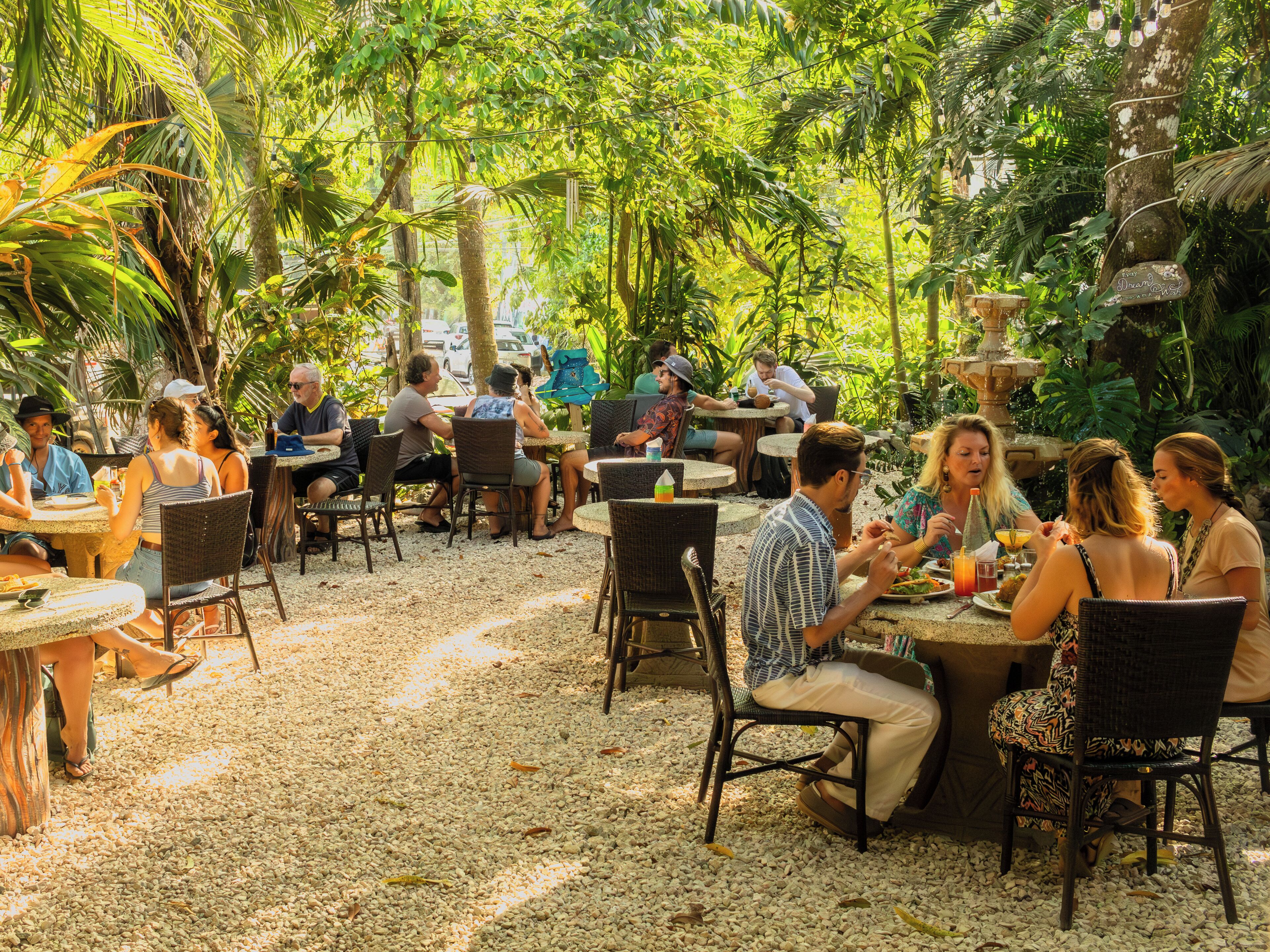 An outdoor dining area with round tables and wicker chairs, surrounded by lush greenery and a pebbled ground.