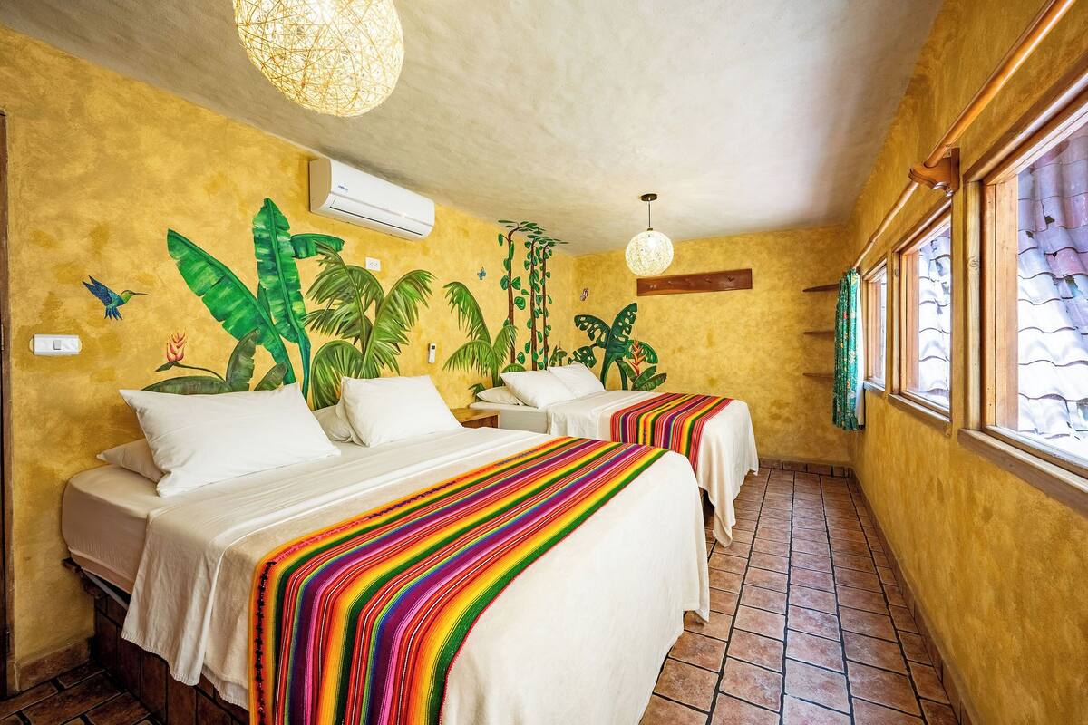 A room with two beds, a mural of tropical plants, a hanging light fixture, and a window with curtains.