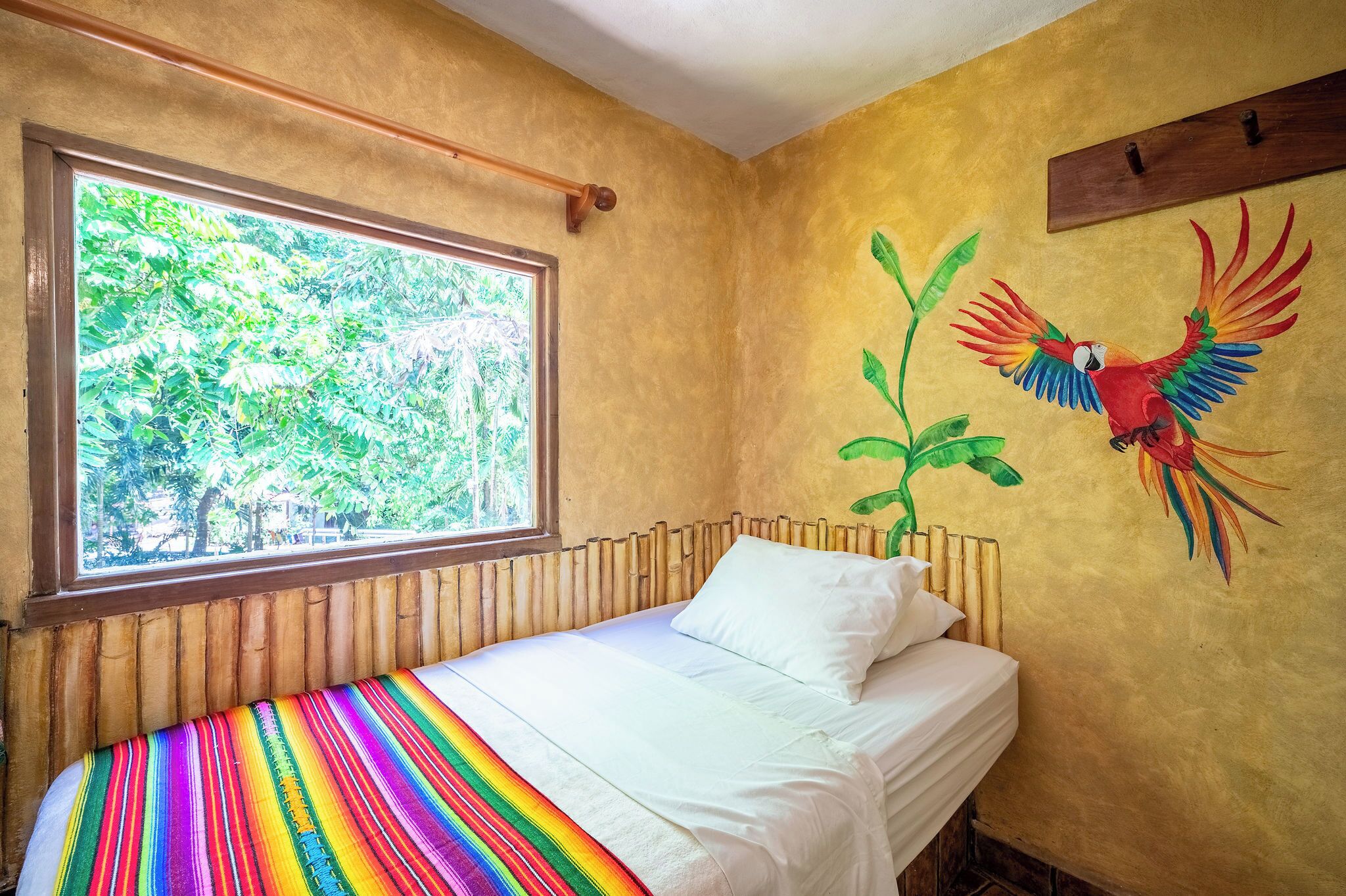 A room with a bed, a window, and a mural of a parrot.