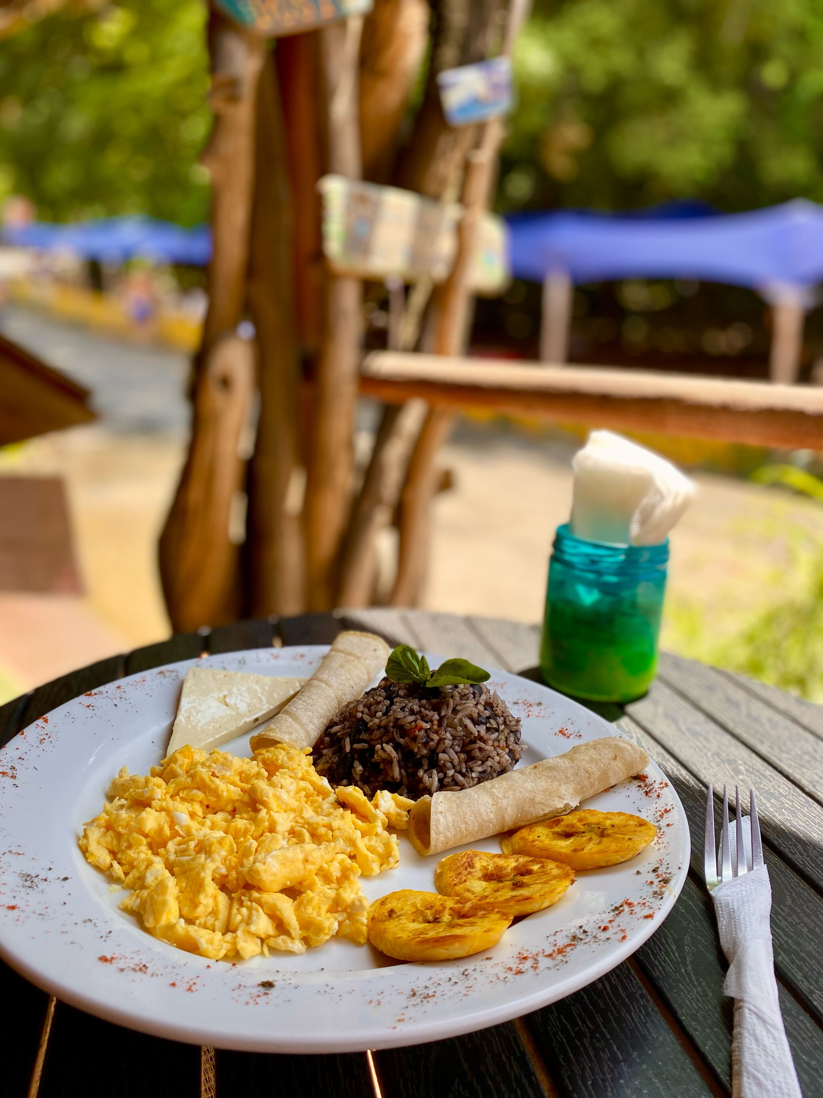 Une assiette de nourriture comprenant des œufs brouillés, du riz noir, des bananes plantains et une tortilla, le tout servi dans une assiette blanche.
