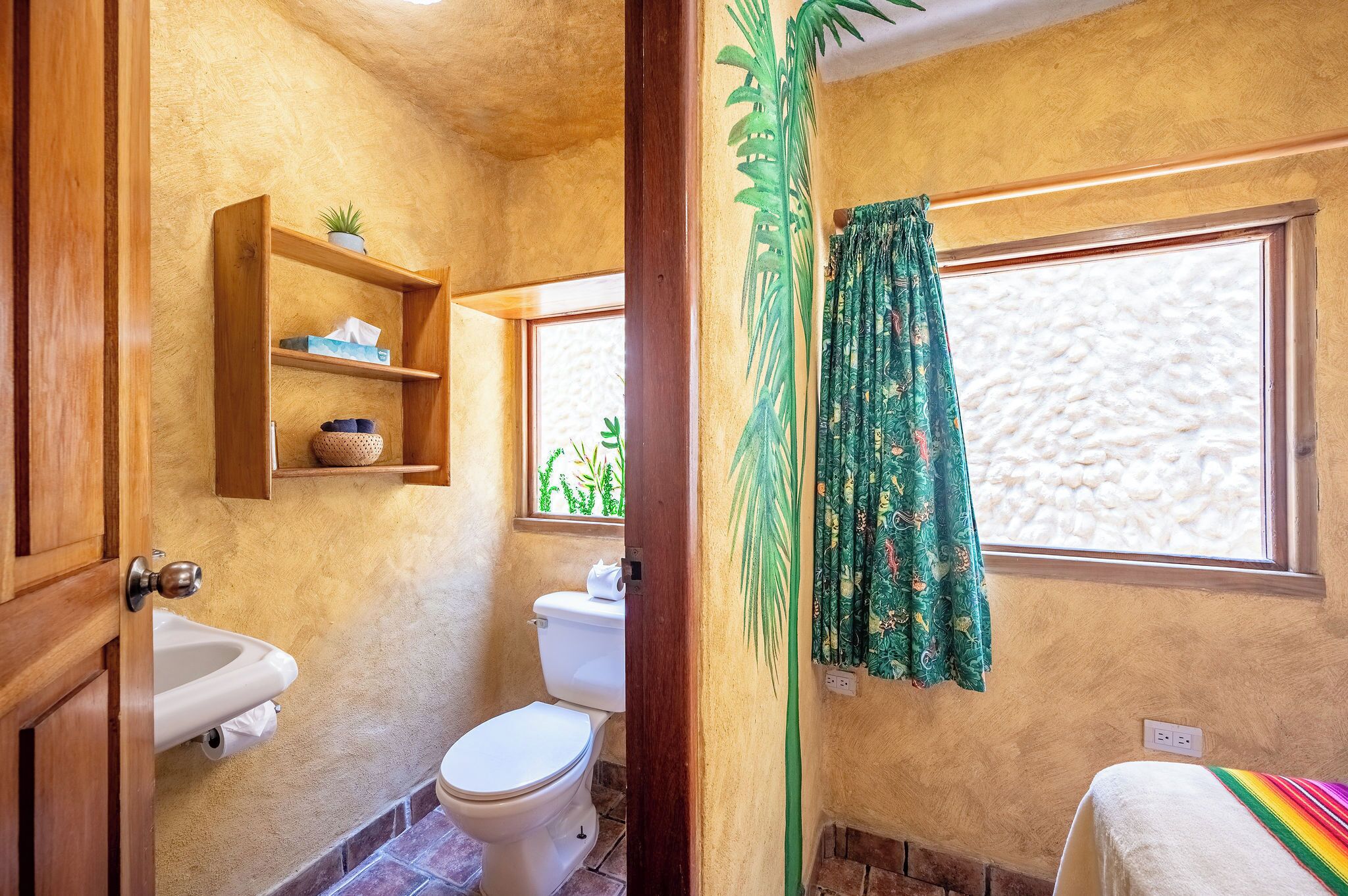 A bathroom with a toilet, sink, shelf with a plant, and a window with curtains.