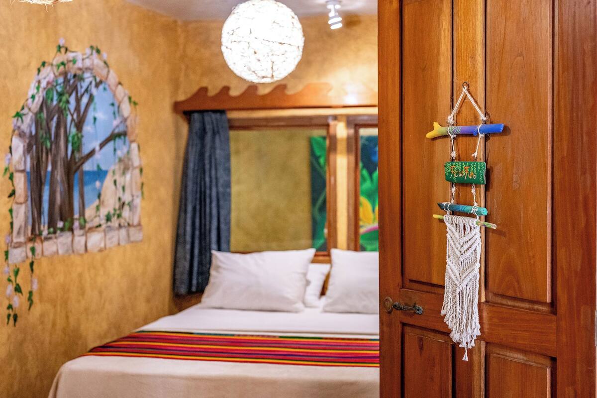 A bedroom with a bed, a wooden door, a wall painting, and a hanging macrame decoration.