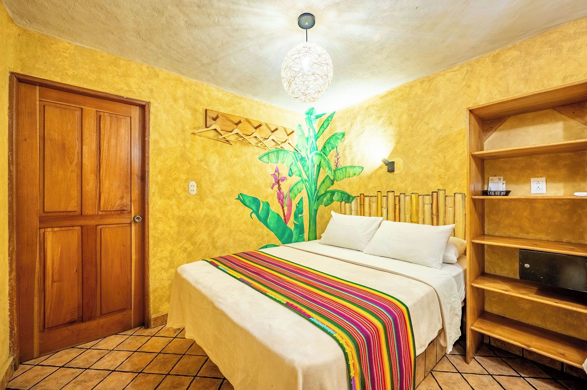 A room with a bed, a wooden door, a wall-mounted shelf, and a ceiling light fixture.