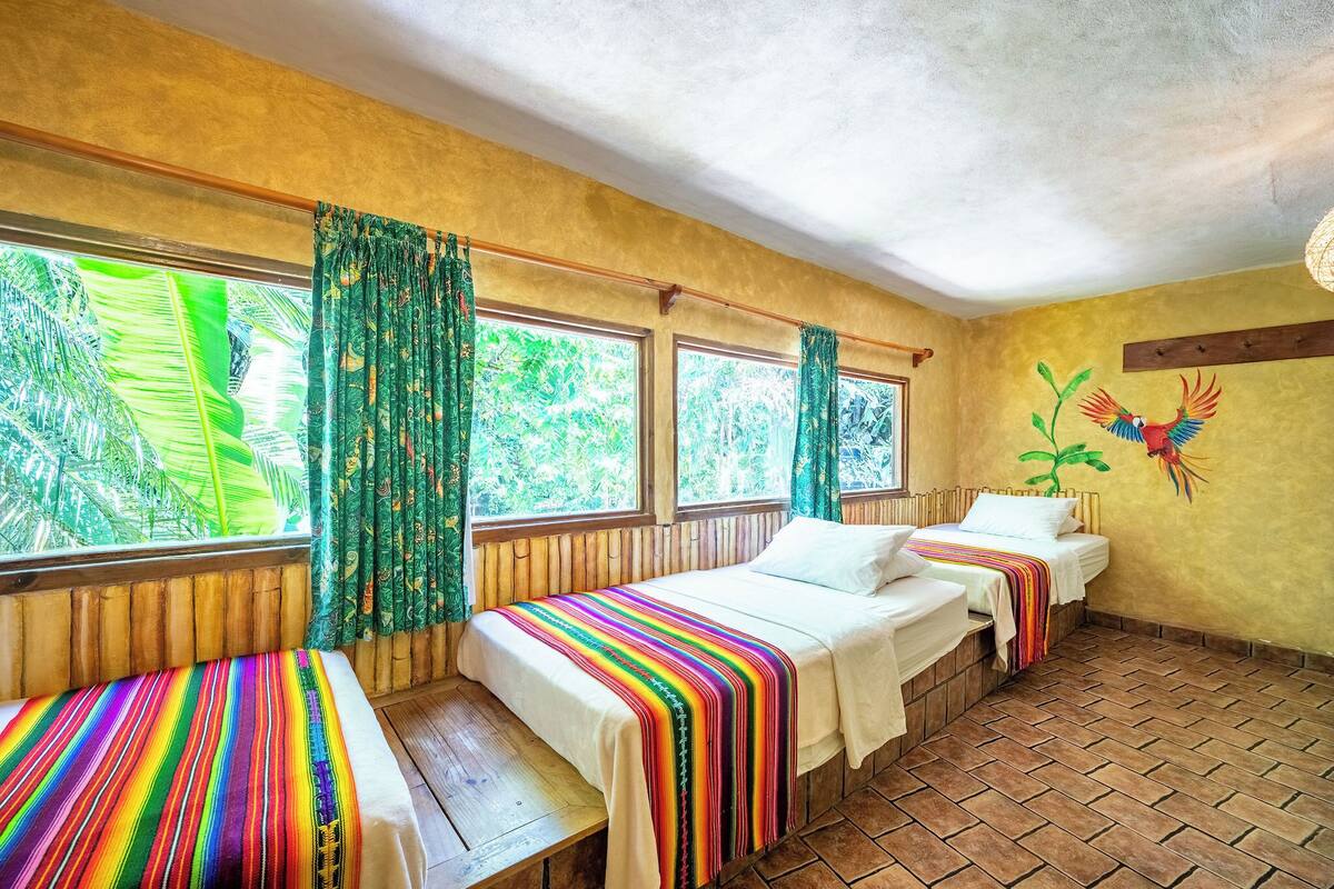 A room with two beds, a bench, and large windows with curtains.