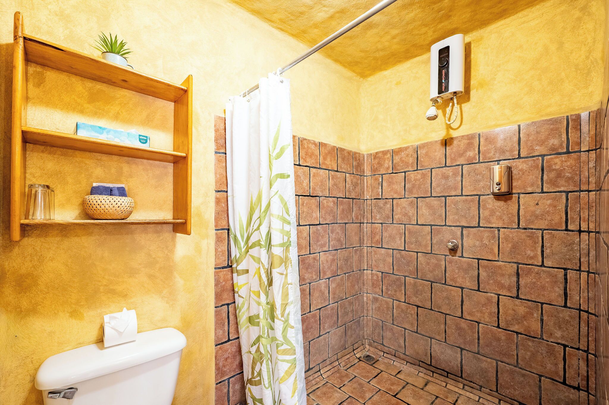 Ein Badezimmer mit Toilette, einer Dusche mit Bambusvorhang, Ablagefläche für Toilettenartikel und einem Wanddurchlauferhitzer.
