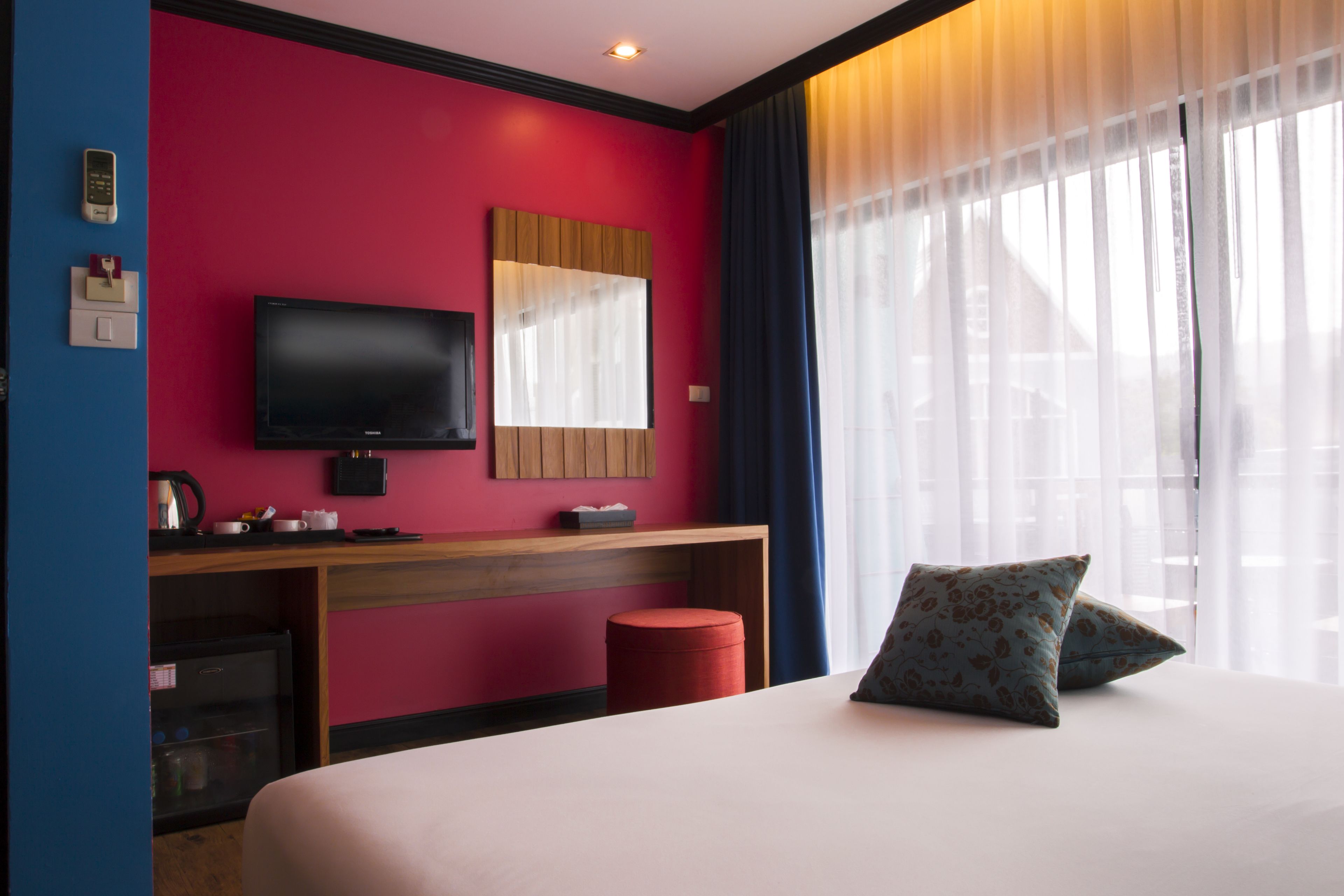 A hotel room with a large bed, a TV mounted on the wall, a wooden desk, and a red ottoman.