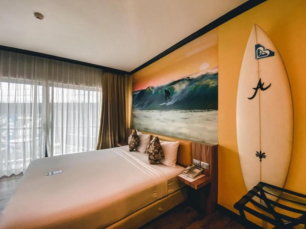 Una habitación de hotel con una cama, una mesita de noche, una tabla de surf y un cuadro de un surfista sobre una ola.