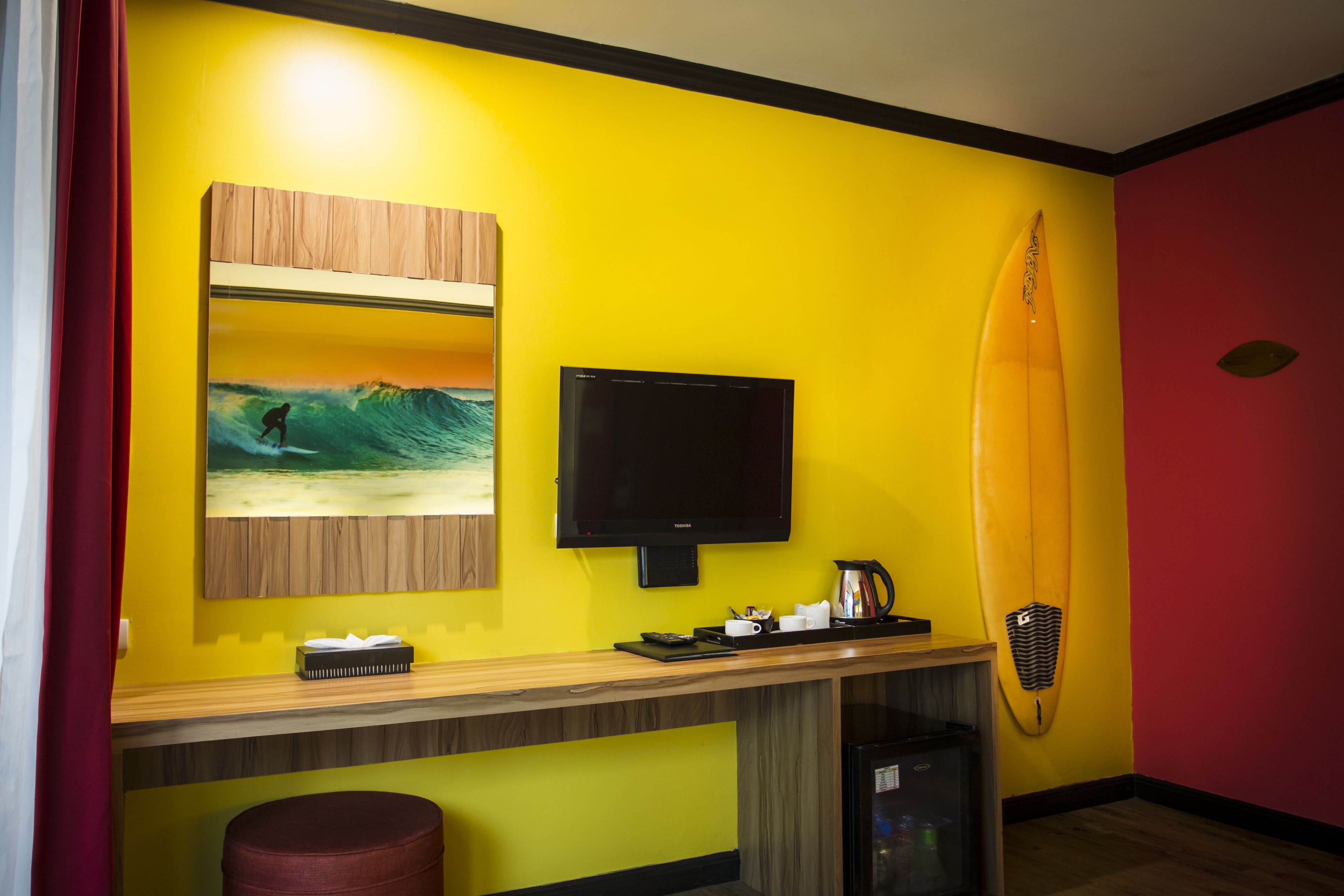 A room with a yellow wall, a mounted TV, a surfboard, a wooden counter, and a red curtain.