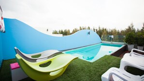 Un área de piscina con tobogán, césped artificial y sillas de descanso.