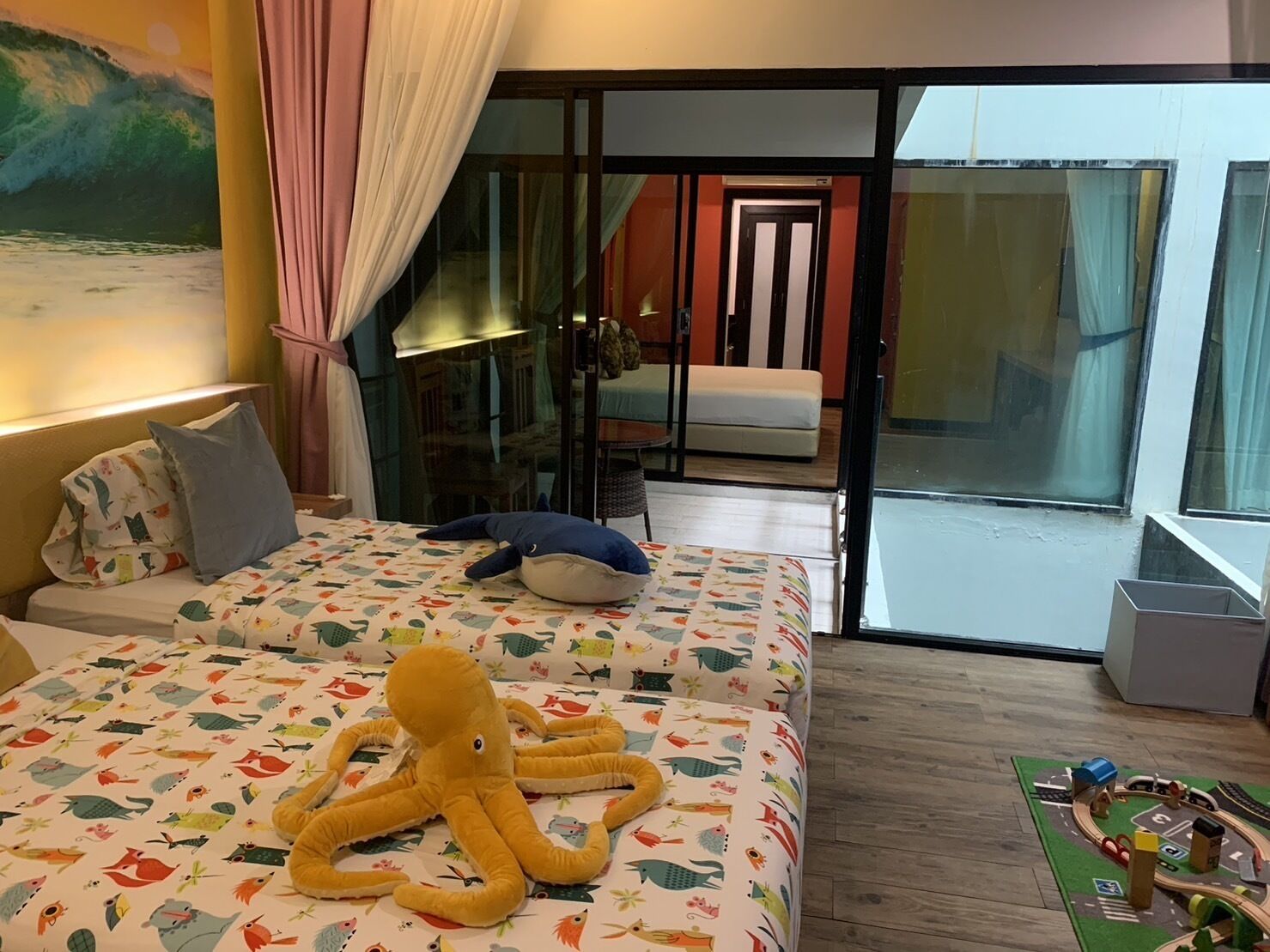 A hotel room with a bed featuring a colorful octopus plush toy, a view of the outdoor area, and a toy train set on the floor.