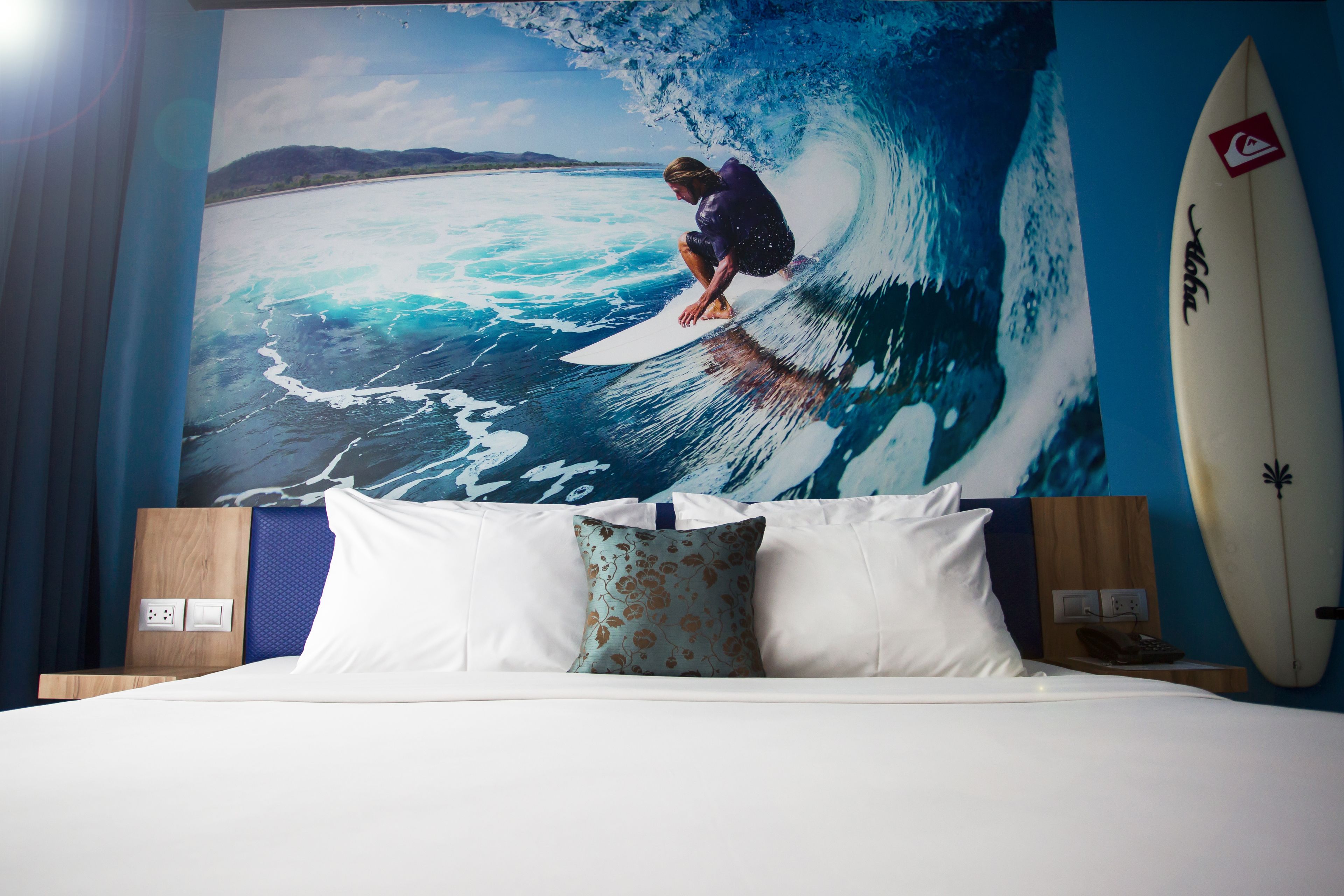A hotel room with a large mural of a surfer on a wave, a bed with white and blue pillows, and a surfboard leaning against the wall.