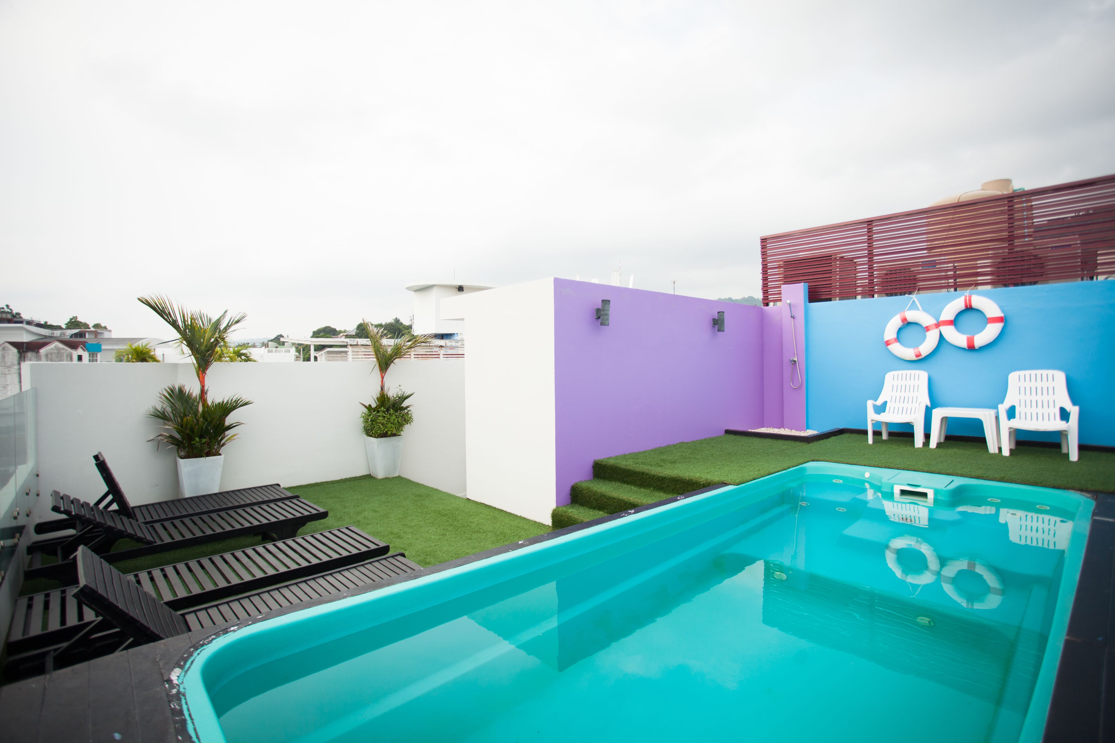 A swimming pool with a clear blue water, surrounded by white walls and a purple wall. There are two white lounge chairs and a white chair on the right side of the pool.