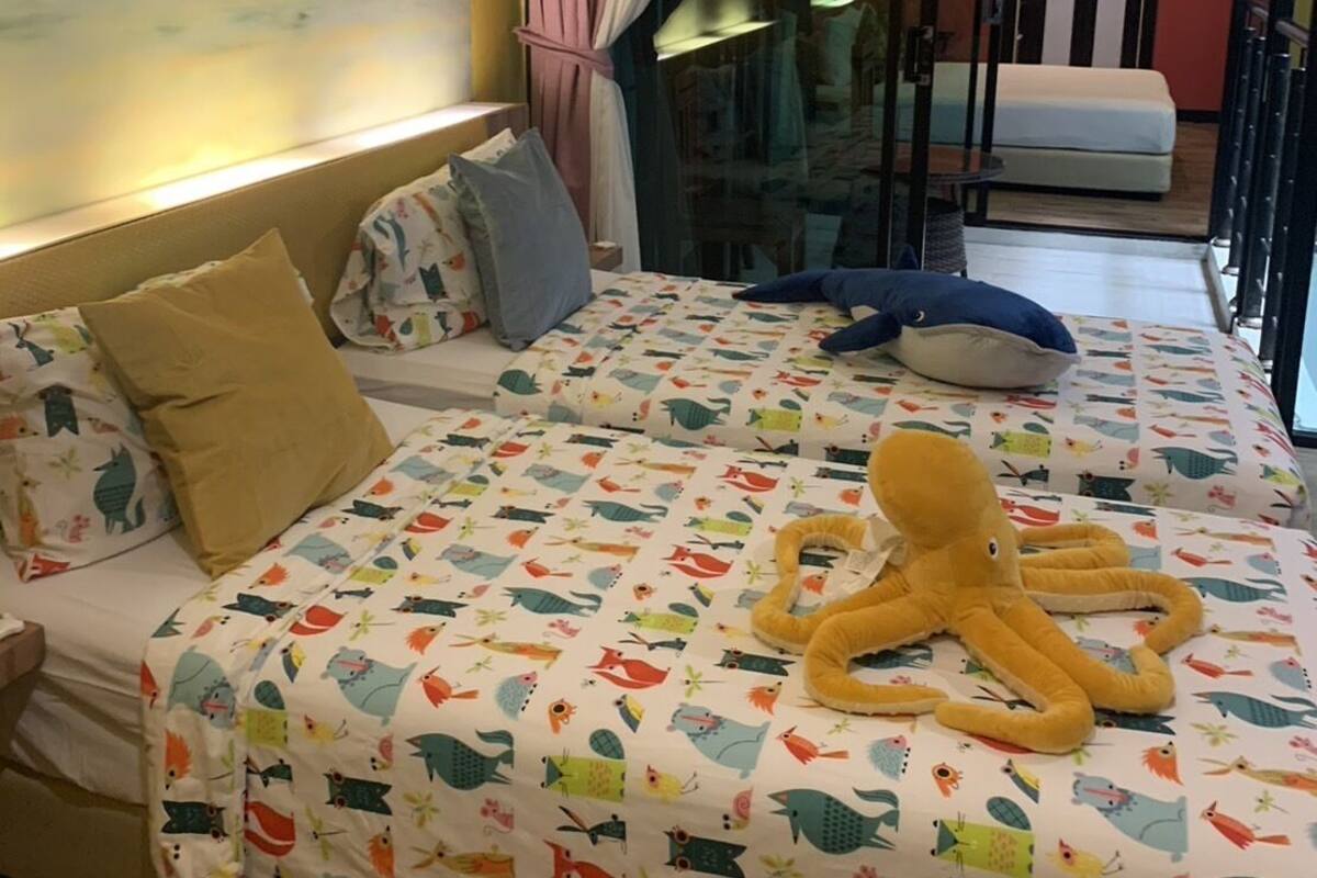 Une chambre à coucher avec un lit recouvert d’une couette aux motifs animaliers colorés, un gros jouet en forme de poulpe et une fresque murale représentant un surfeur sur une vague.