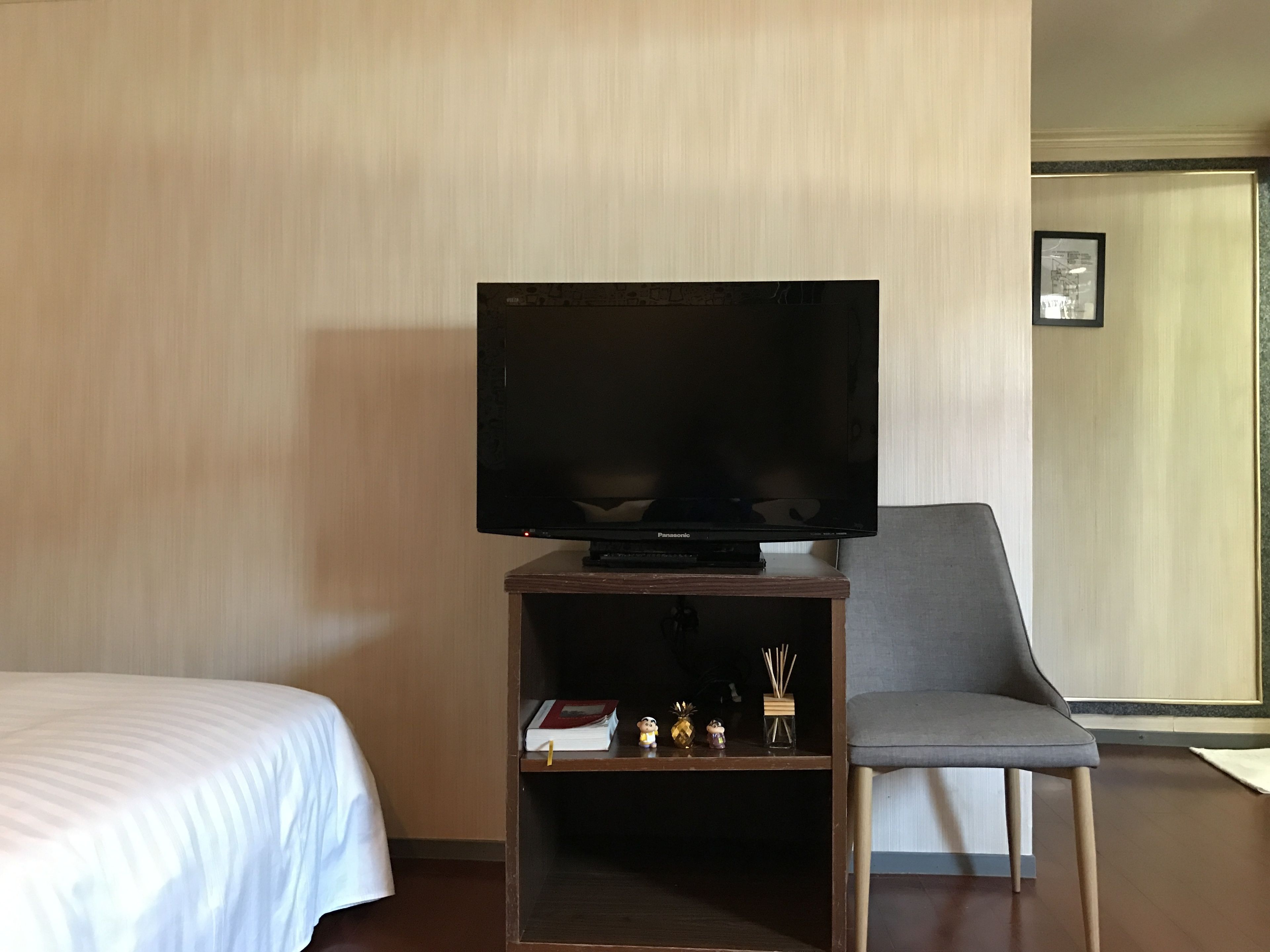 A hotel room with a bed, a television on a wooden stand, a grey armchair, and a small shelf with books and decorative items.