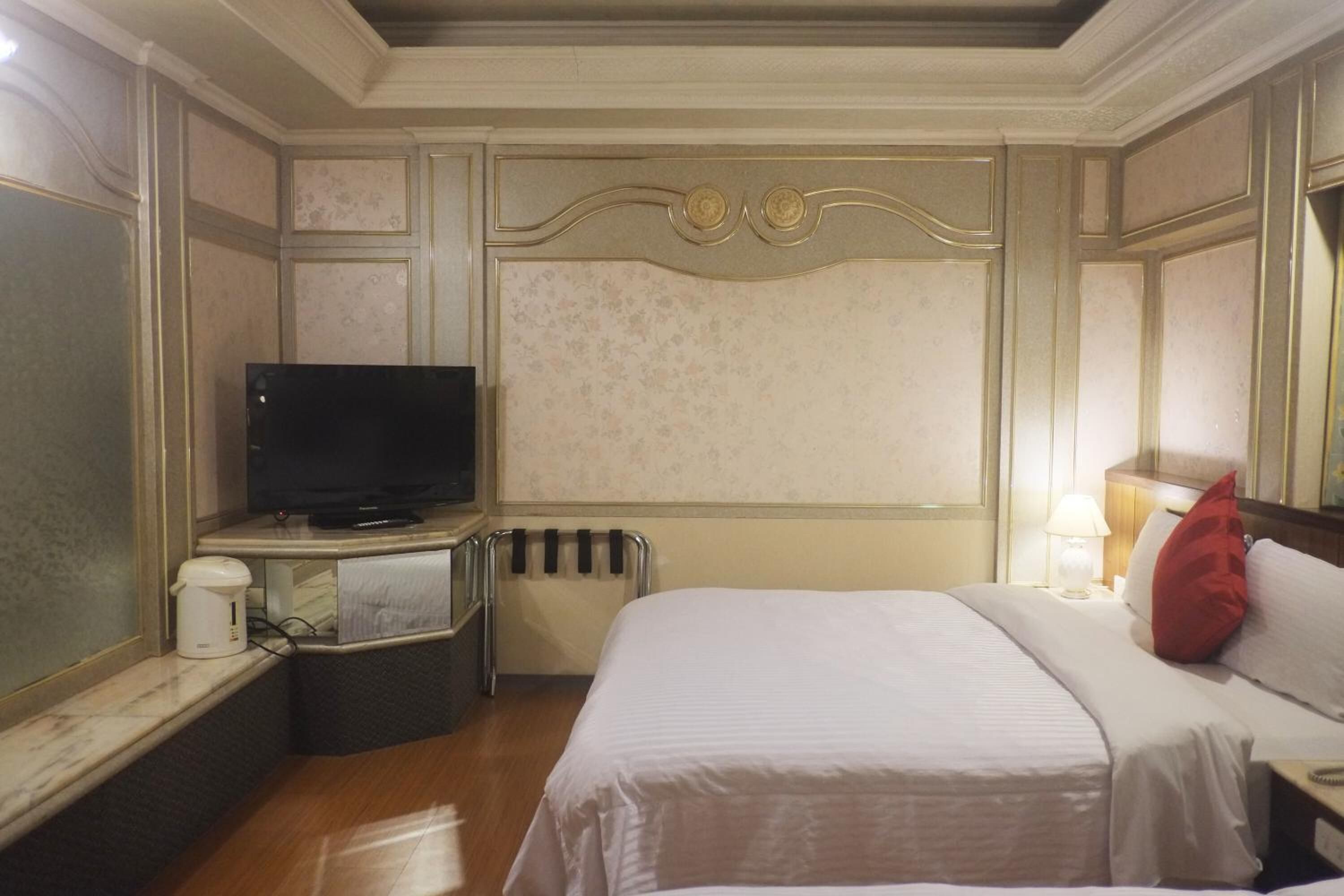 A hotel room with a bed, a television, a bedside table with a lamp, and a radiator.