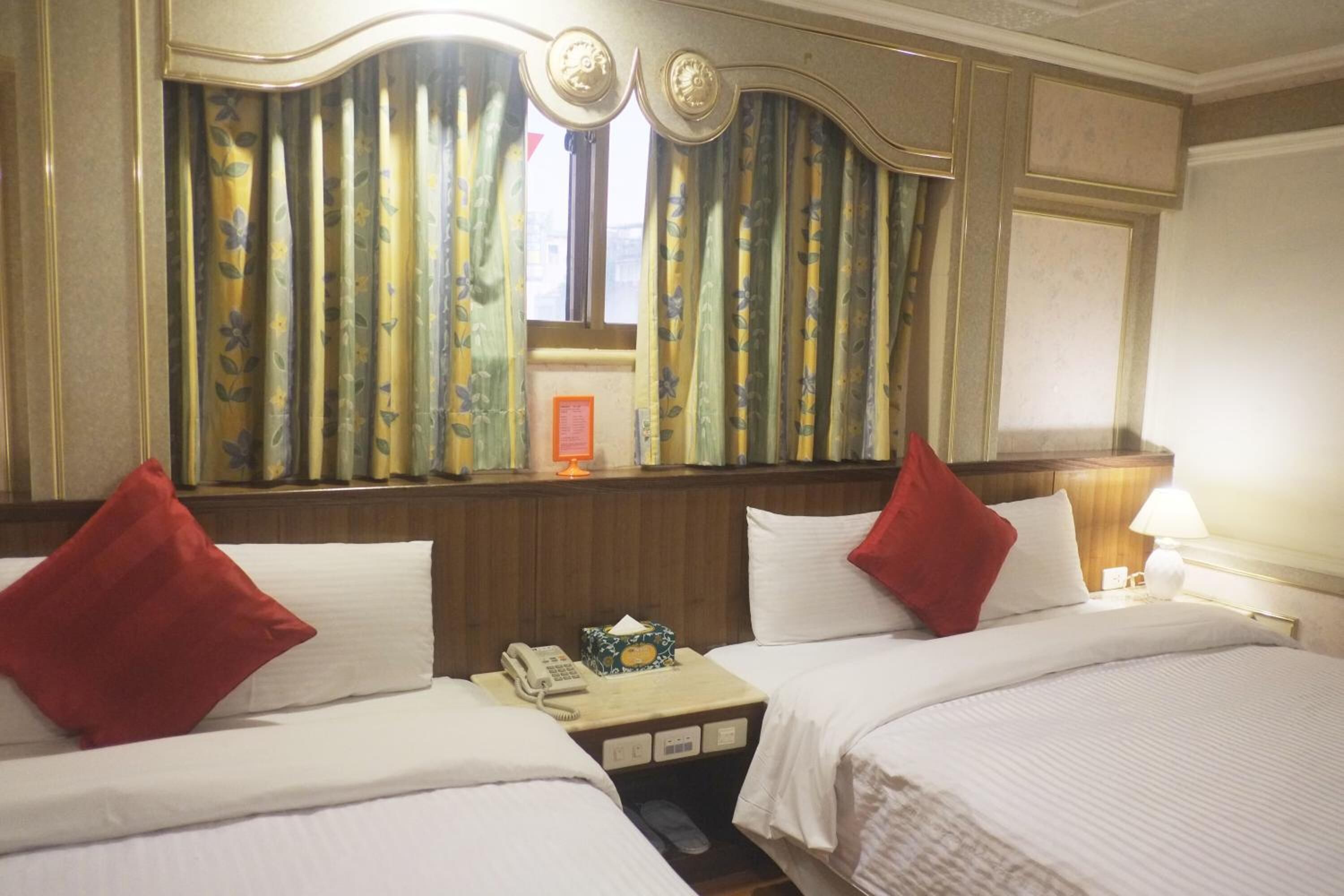 Una habitación de hotel con dos camas, una mesita de noche con teléfono y pañuelos, una lámpara en la mesita y una ventana con cortinas.