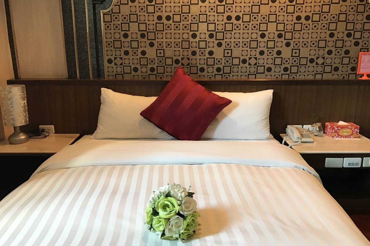 A hotel room with a bed, a nightstand with a lamp, a telephone, and a small table with a box.