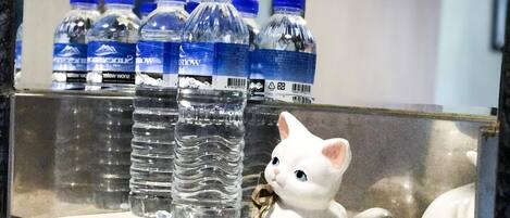 Une salle de bain avec un lavabo, un miroir et une étagère sur laquelle se trouvent des bouteilles d’eau et une statuette de chat blanc.