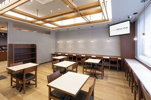 Un espace repas moderne avec des tables et des chaises en bois, un grand écran affichant le nom « SOTETSU FRÉSA INN », et un plafond orné de poutres en bois et doté d’un éclairage encastré.