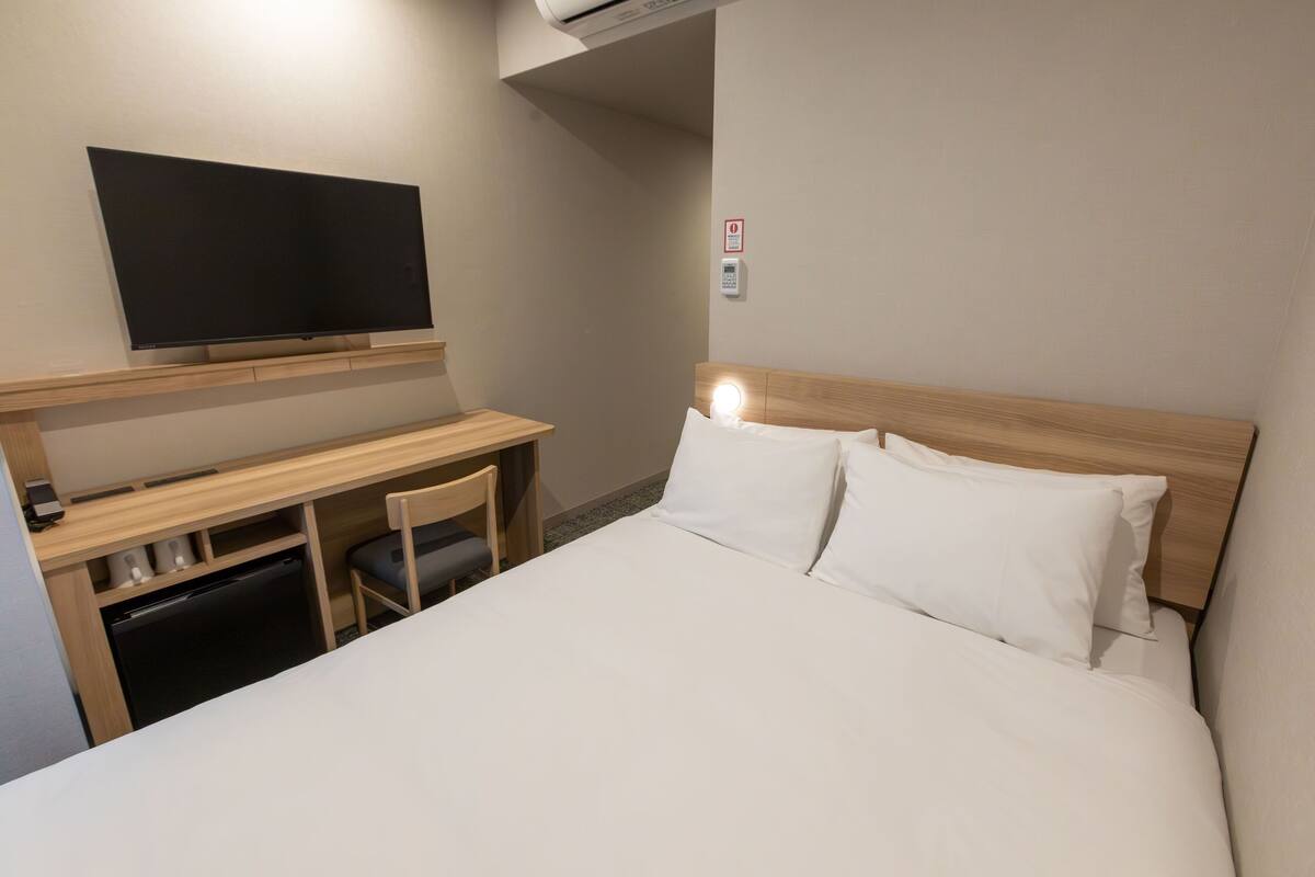 A hotel room with a bed, a desk, a chair, a television, and a wall-mounted control panel.