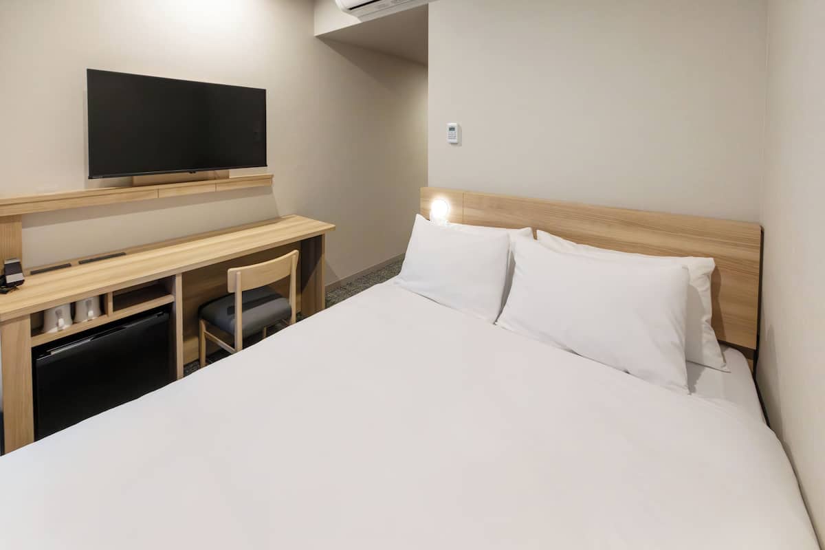 A hotel room with a bed, a desk, a chair, a television, and a wall-mounted control panel.