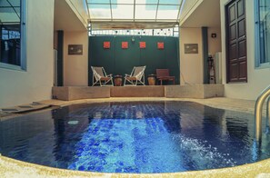 An indoor swimming pool with a clear blue water surface, surrounded by stone steps and a covered seating area with two chairs and a bench. - Boondaree Home Resort (Karon)