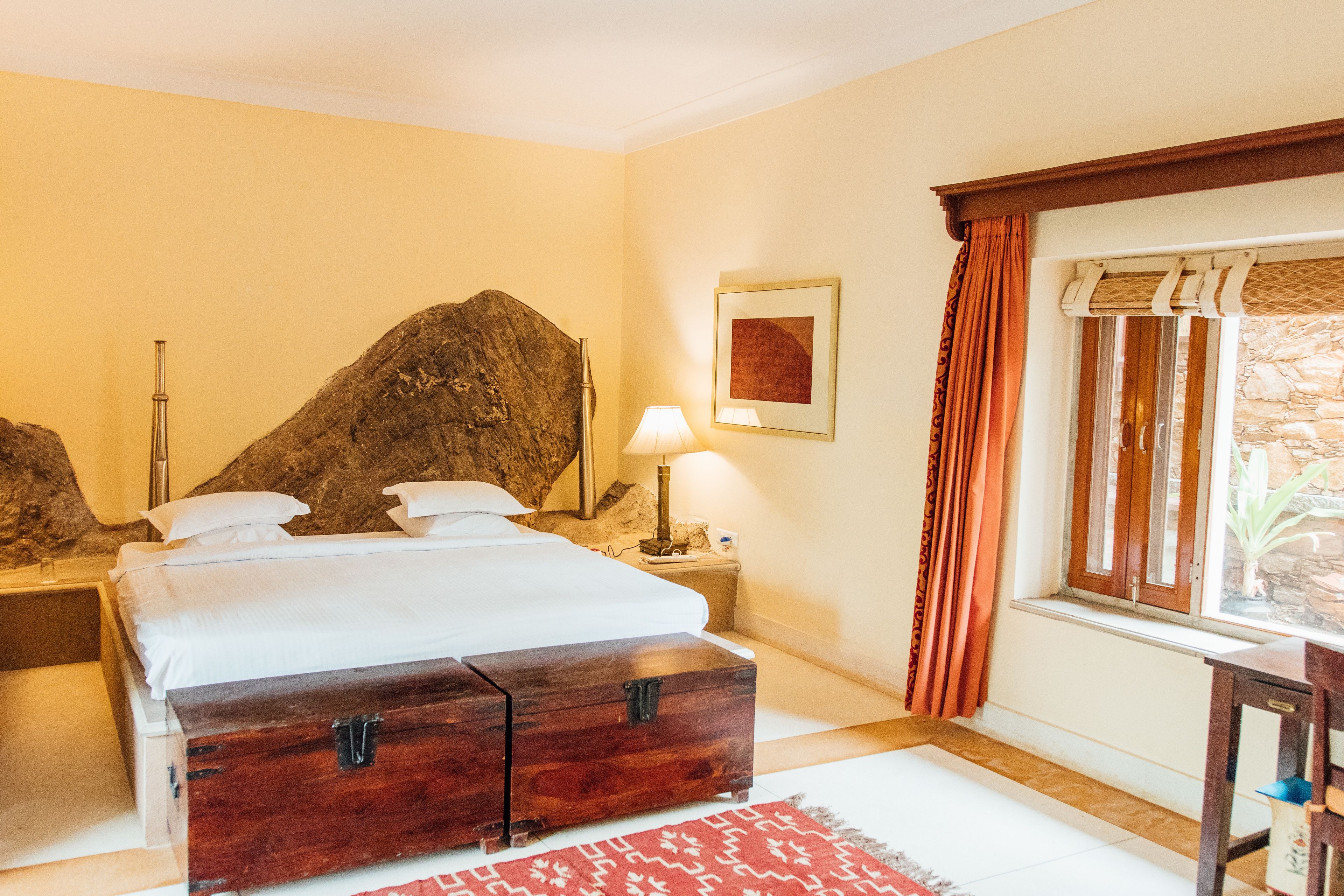 Een hotelkamer met een bed, een nachtkastje, een lamp, een grote rotsdecoratie, een raam met gordijnen en een ingelijst schilderij aan de muur.
