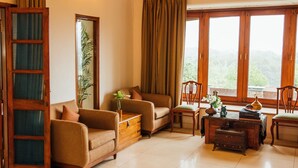 Una sala de estar acogedora con un sofá, sillas, una mesa de centro de madera y vistas a la vegetación a través de amplios ventanales.