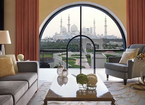 A room with a large arched window, a sofa, a chair, a coffee table with decorative items, and a view of a white building with domes and minarets. - The Ritz-Carlton Abu Dhabi, Grand Canal (Abu Dhabi)