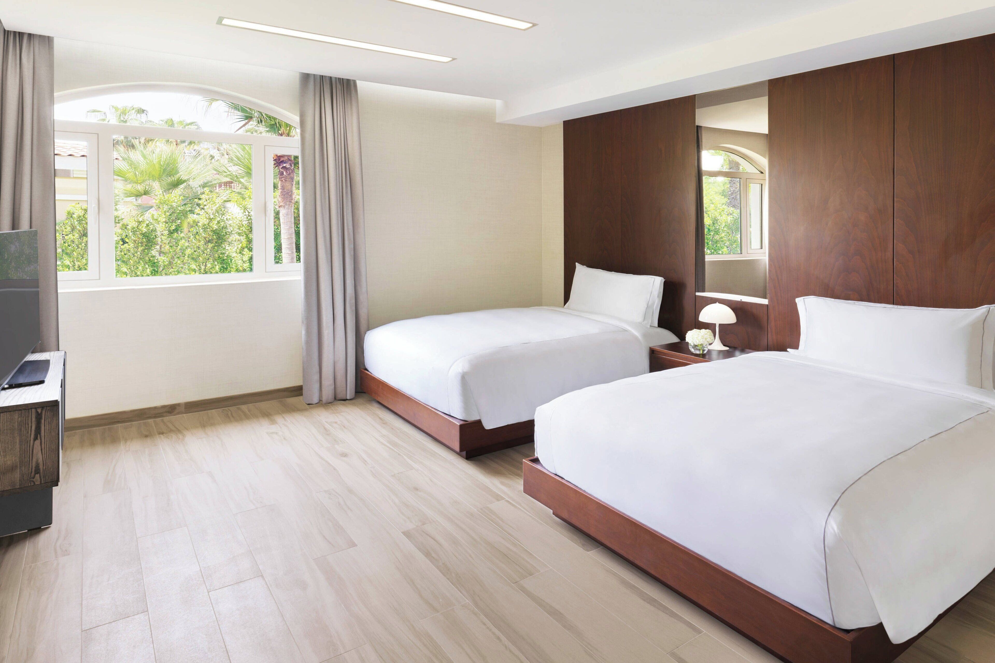 A hotel room with two beds, a TV, a desk, and a window with curtains.