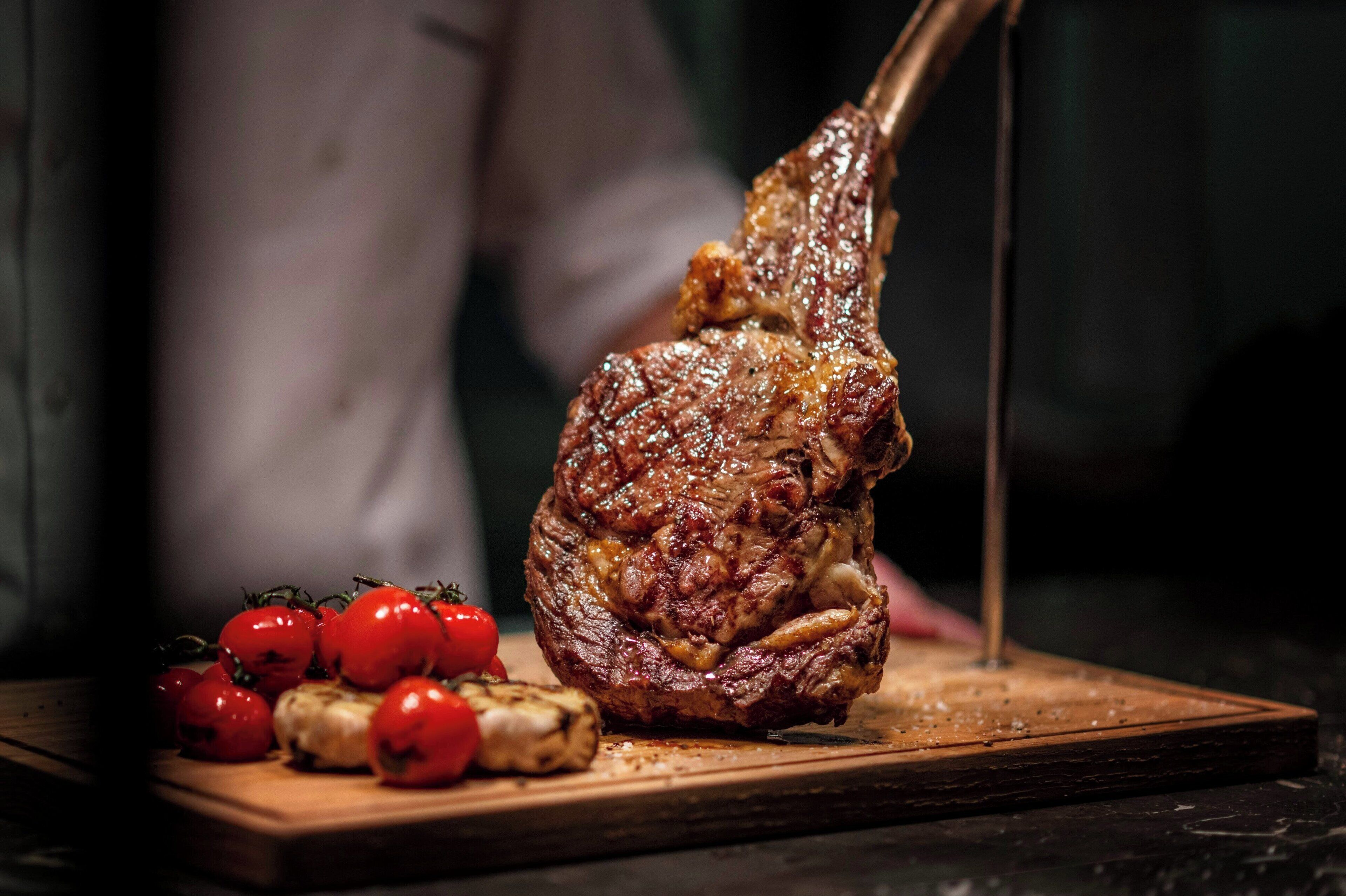 A cooked steak on a wooden board with cherry tomatoes and garlic cloves beside it.
