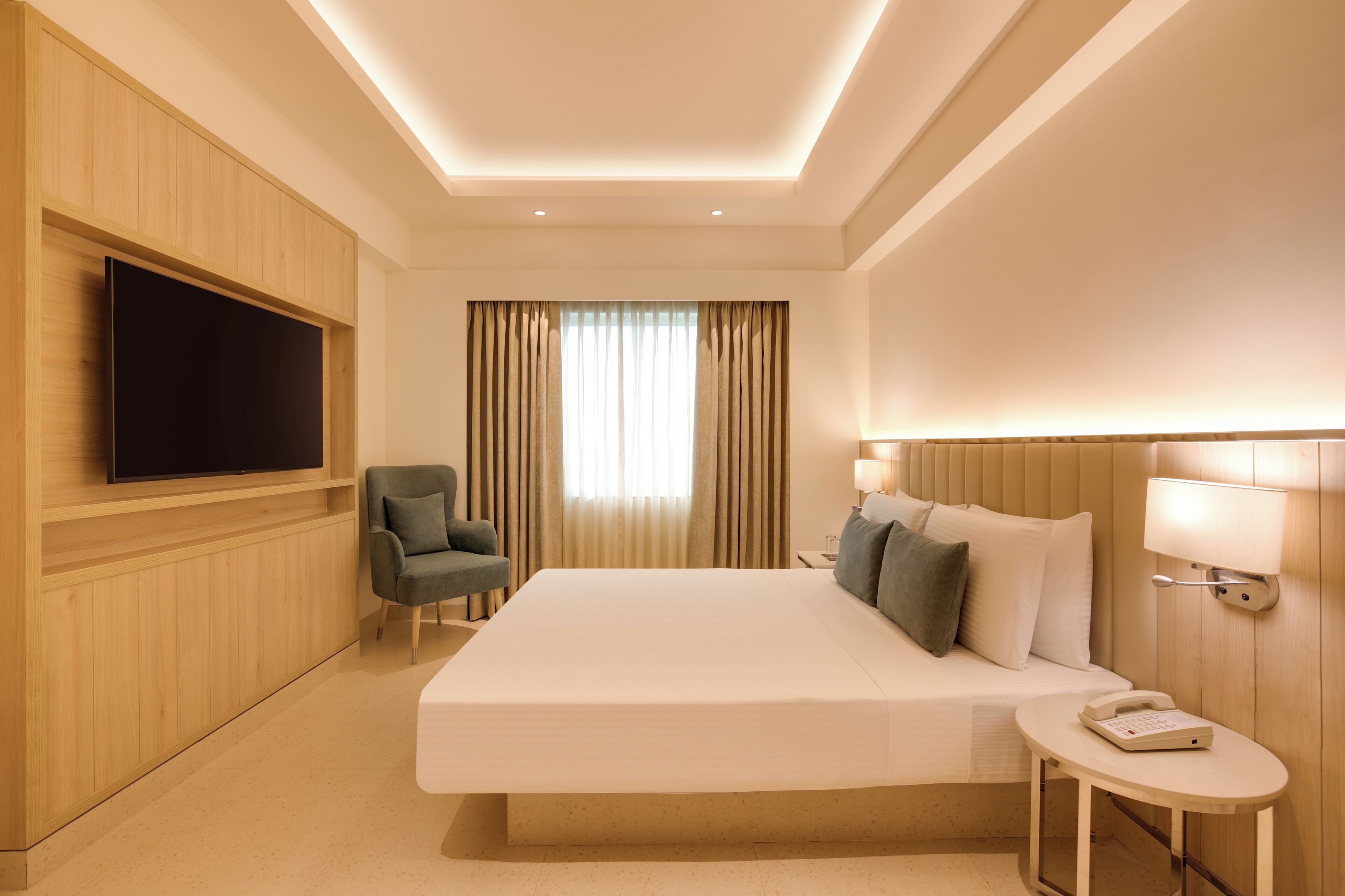 A hotel room with a large bed, a television mounted on the wall, a chair, a bedside table with a lamp, and a phone.