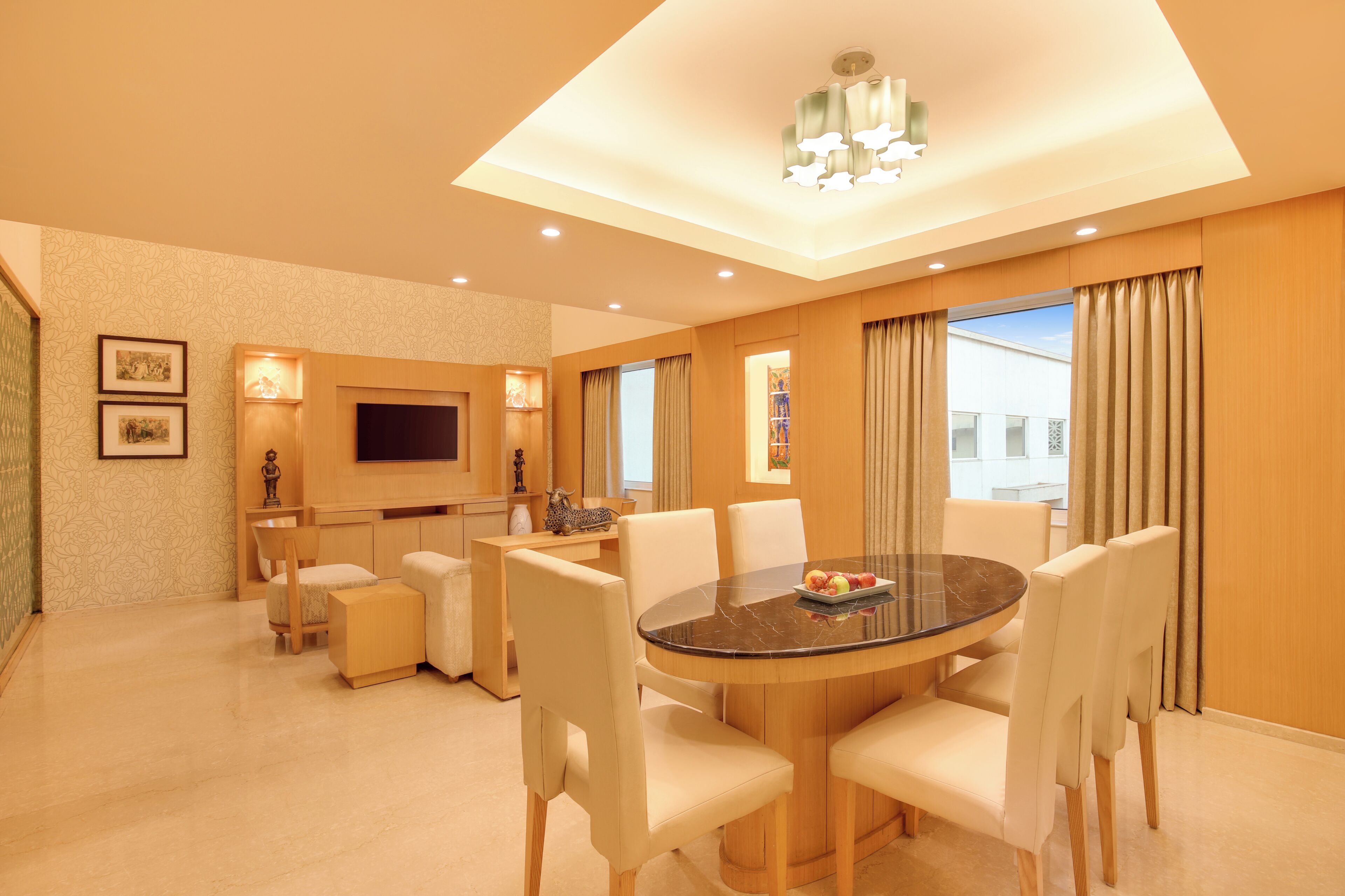 Habitación de hotel con una mesa de comedor redonda, sillas blancas, un televisor de pantalla plana y un amplio ventanal con cortinas.