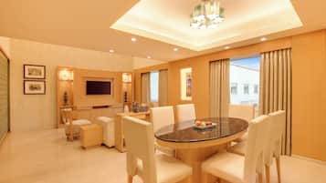 Habitación de hotel con una mesa de comedor redonda, sillas blancas, un televisor de pantalla plana y un amplio ventanal con cortinas.
