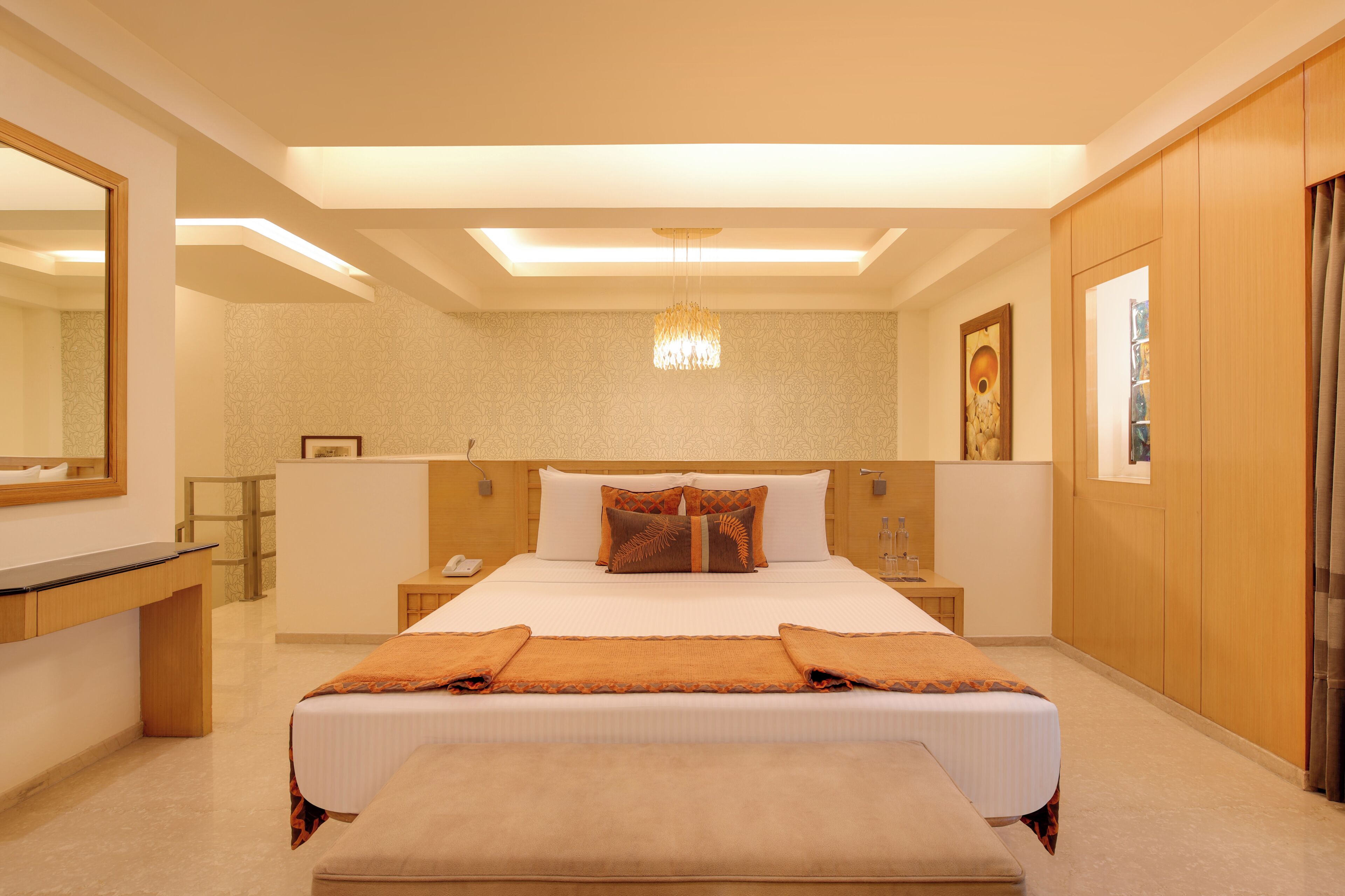 A hotel room with a large bed, bedside tables, a desk, a mirror, a chandelier, and artwork on the walls.