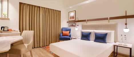 A hotel room with a large bed, a desk with a chair, a small table with a lamp, and a window with curtains.