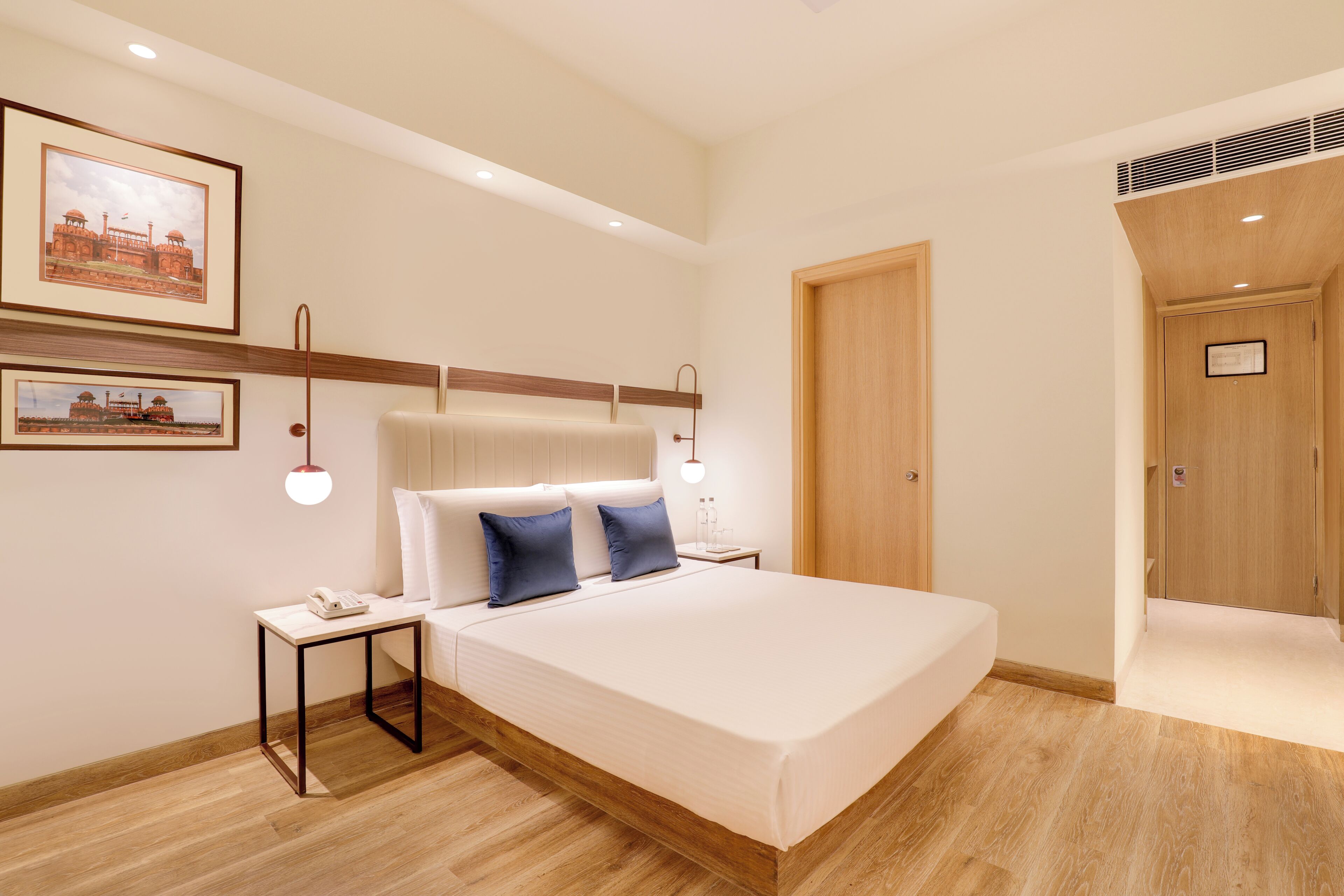 A modern hotel room with a large bed, bedside tables, a nightstand, framed pictures on the wall, and a wooden door.