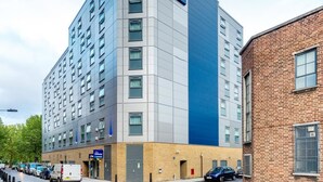 A modern Travelodge hotel with a prominent blue and grey facade, multiple floors, and a brick building adjacent to it.