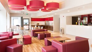 A modern dining area with red upholstered booths and tables, a kitchen area, and a bar with a red accent wall. - Travelodge Liverpool Central Exchange Street (Liverpool)
