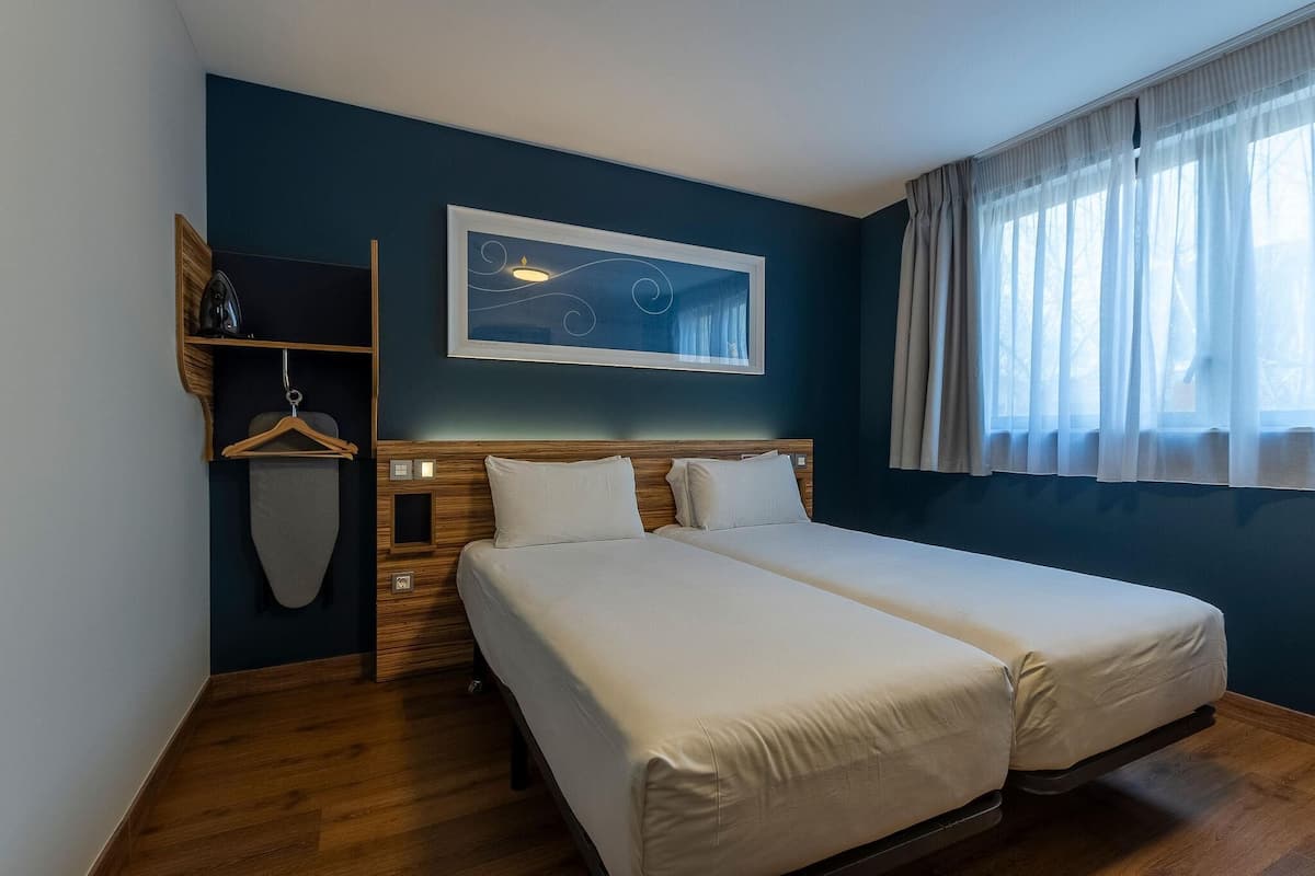 Hotellihuone, jossa on kaksi sänkyä, puinen pääty, seinälle kiinnitetty taideteos, ikkuna, jossa on läpikuultavat verhot, sekä puinen hylly, jossa on pyyhe ja henkari.