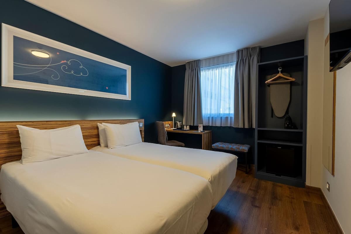 Hotellihuone, jossa on kaksi sänkyä, työpöytä, tuoli, vaatekaappi ja suuri abstrakti maalaus seinällä.