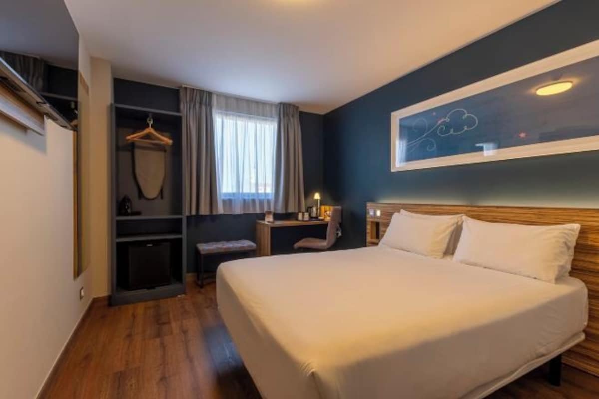 Une chambre d’hôtel avec un grand lit, un bureau, une chaise, une armoire et une fenêtre avec des rideaux.