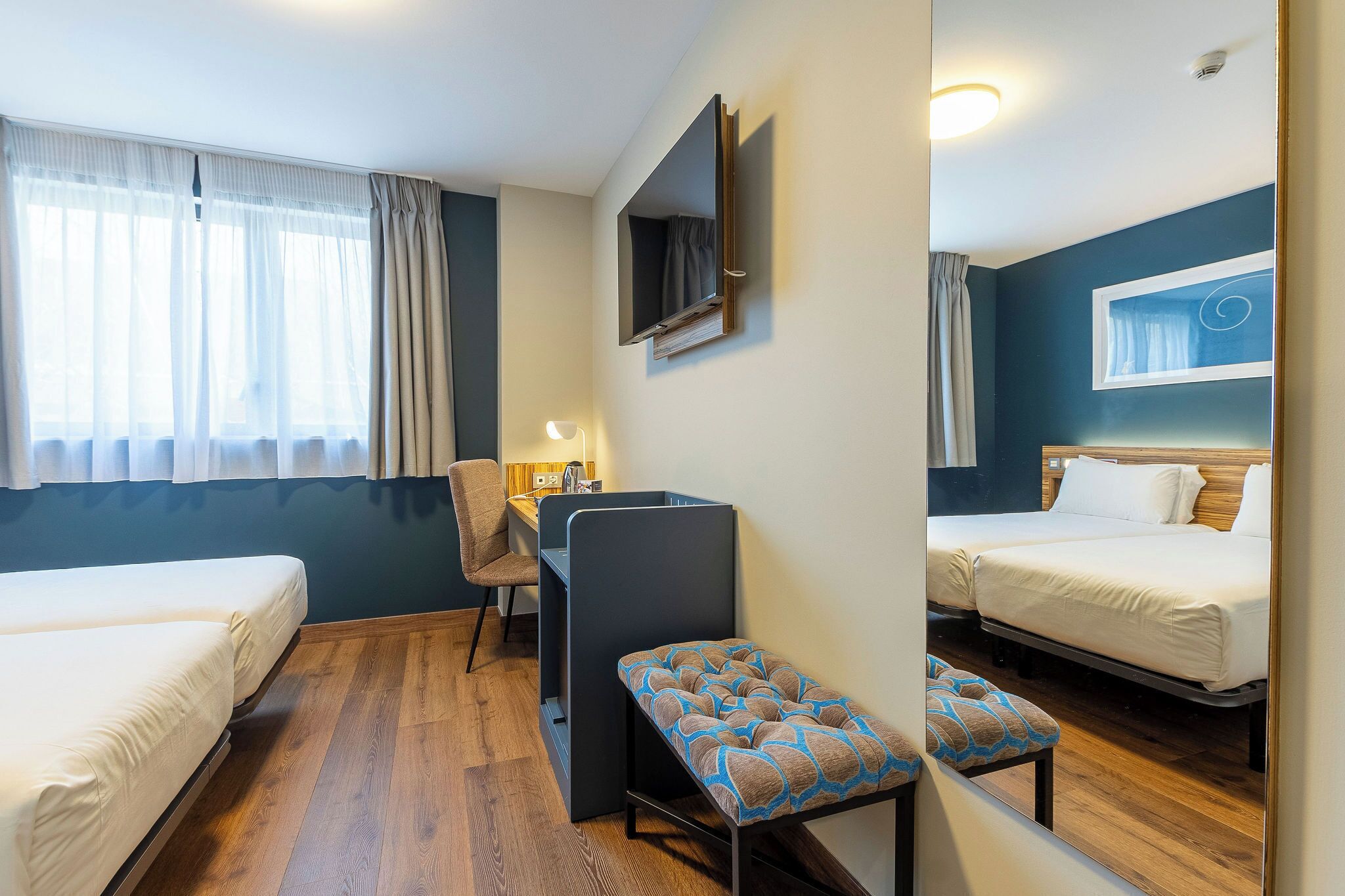 Habitación de hotel con dos camas, un escritorio, una silla, un banco y un televisor montado en la pared.
