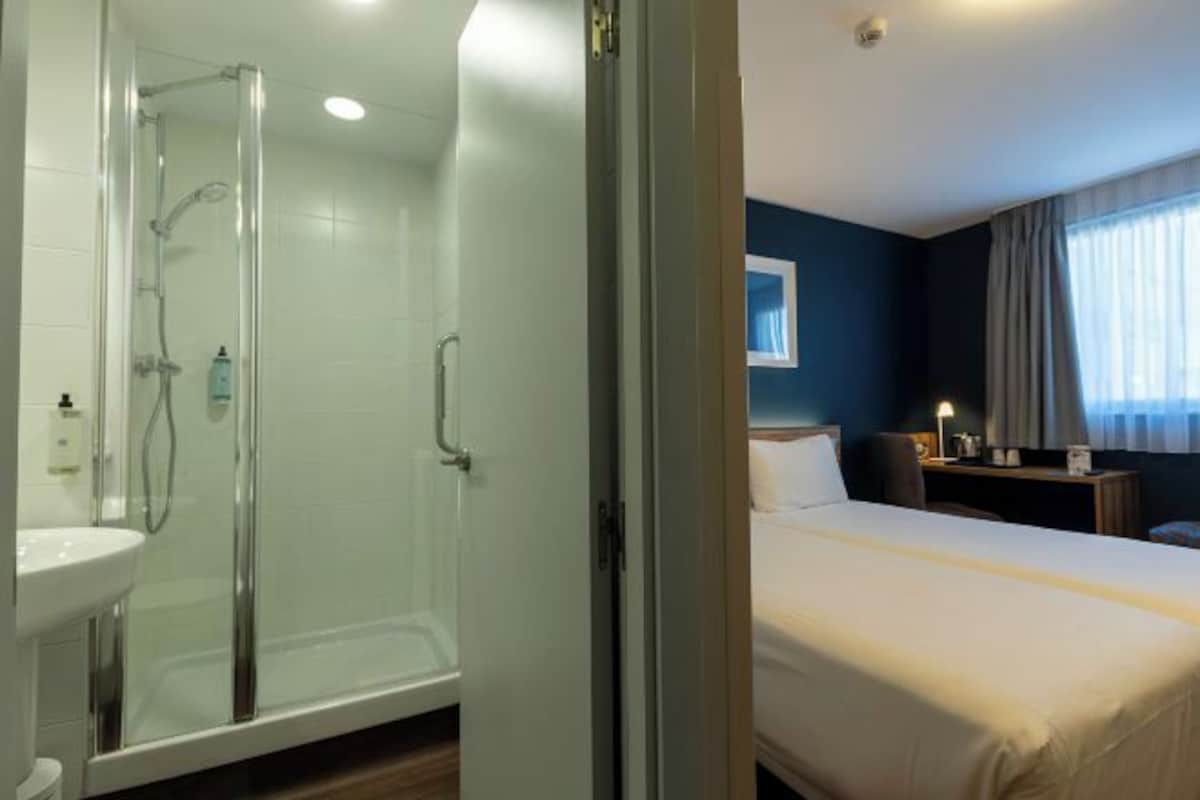 Une chambre d’hôtel avec une douche en verre, un lit, une table de chevet et une fenêtre avec des rideaux.