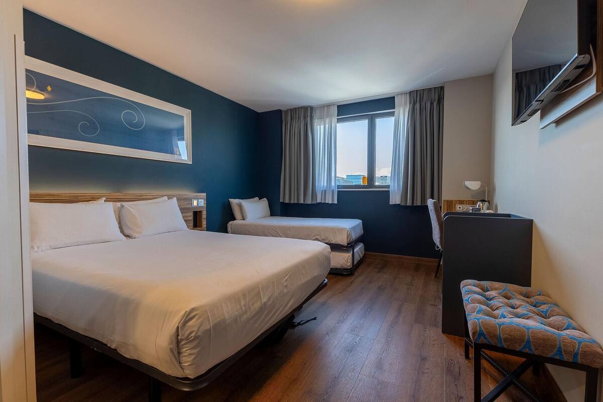 Hotellihuone, jossa on kaksi sänkyä, työpöytä, tuoli, ikkuna verhoilla ja seinään asennettu ilmastointilaite.
