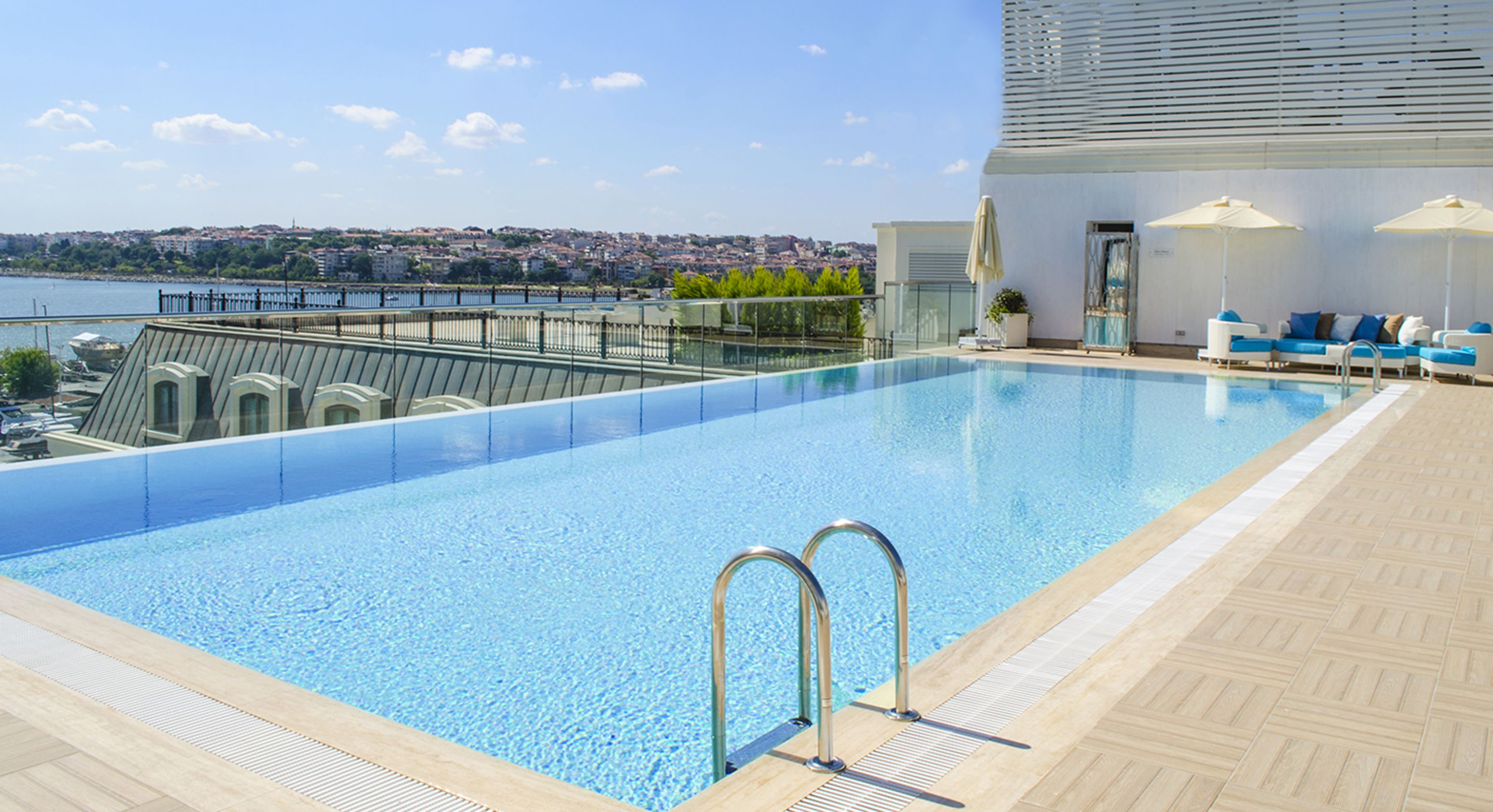 A rooftop pool with clear blue water, a view of a cityscape, and a seating area with umbrellas.
