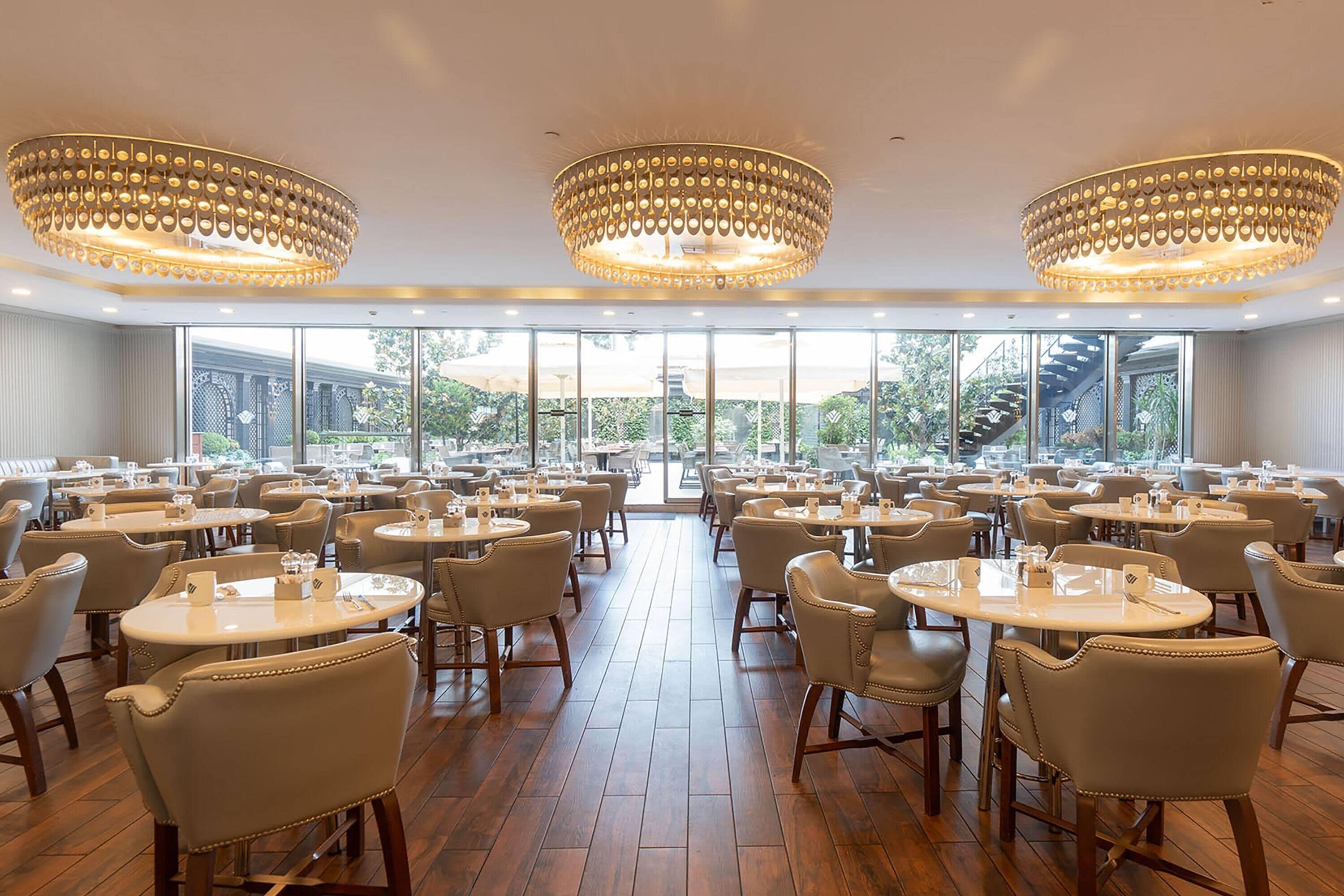 Un vaste espace dédié à la salle à manger, avec des tables et des chaises rondes, de grandes fenêtres et des lustres décoratifs.