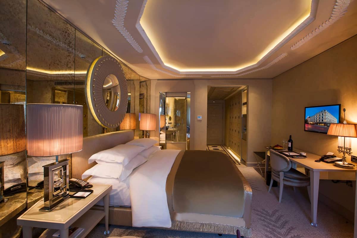 A hotel room with a large bed, bedside lamps, a desk with a computer, and a television on the wall.