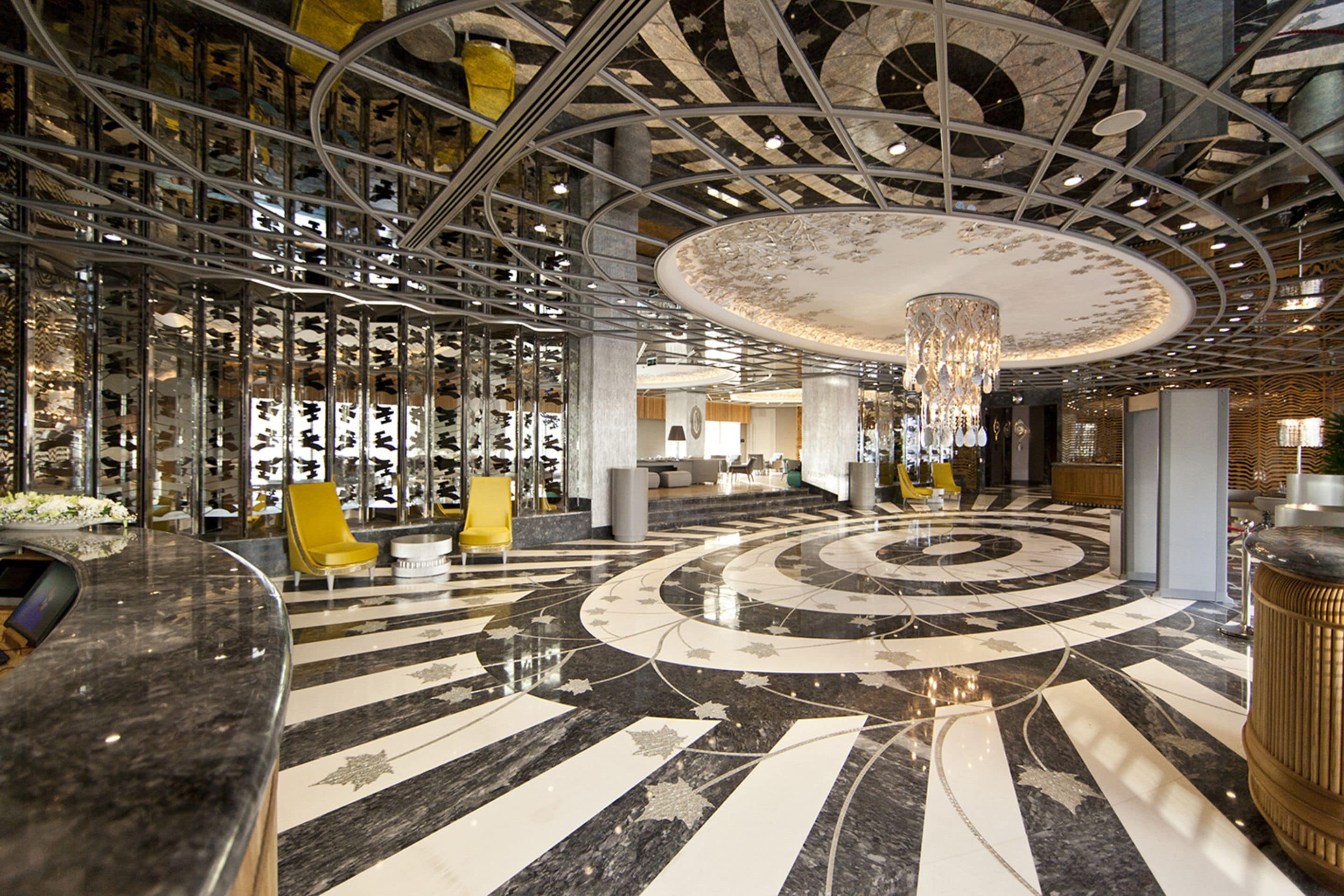 A modern lobby with a circular marble floor, a central chandelier, and a seating area with yellow chairs.